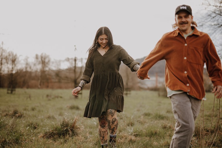 Eine Frau und ein Mann gehen Hand in Hand durch eine Wiese, wunderschön eingefangen von ihrem Fotografen. Die Frau trägt ein dunkelgrünes Kleid und hat Tattoos an den Beinen, während der Mann eine orangefarbene Jacke und eine Baseballkappe trägt. Beide lächeln natürlich, und Bäume und Hügel bieten die perfekte Kulisse für ihr Paarshooting.