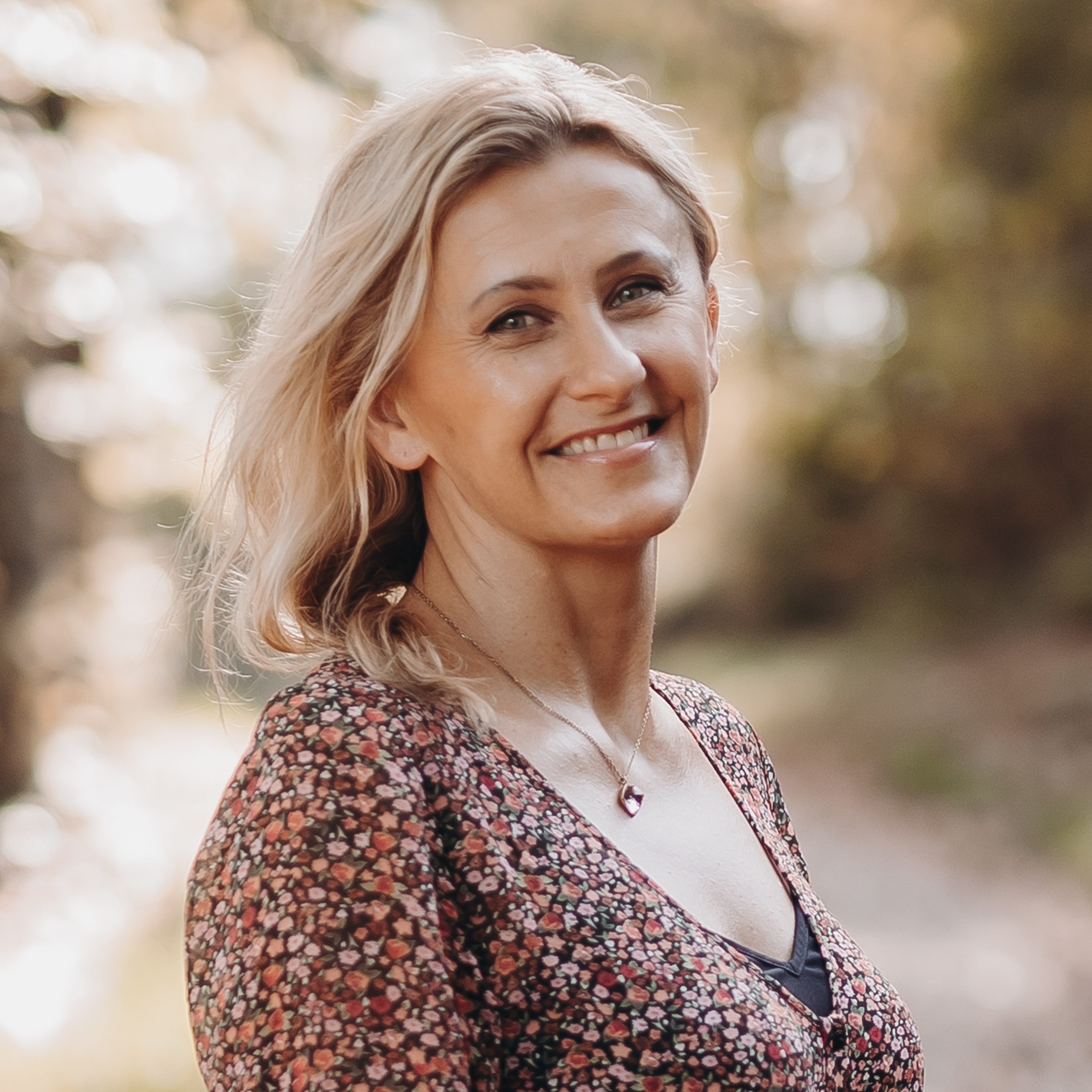 Eine Person mit hellem Haar und einem Oberteil mit Blumenmuster lächelt und blickt in die Kamera bei einem Fotoshooting von Tasha Ender Photography. Der Hintergrund ist verschwommen, mit Andeutungen von Grün und durchscheinendem Sonnenlicht, was eine Szenerie im Freien suggeriert.