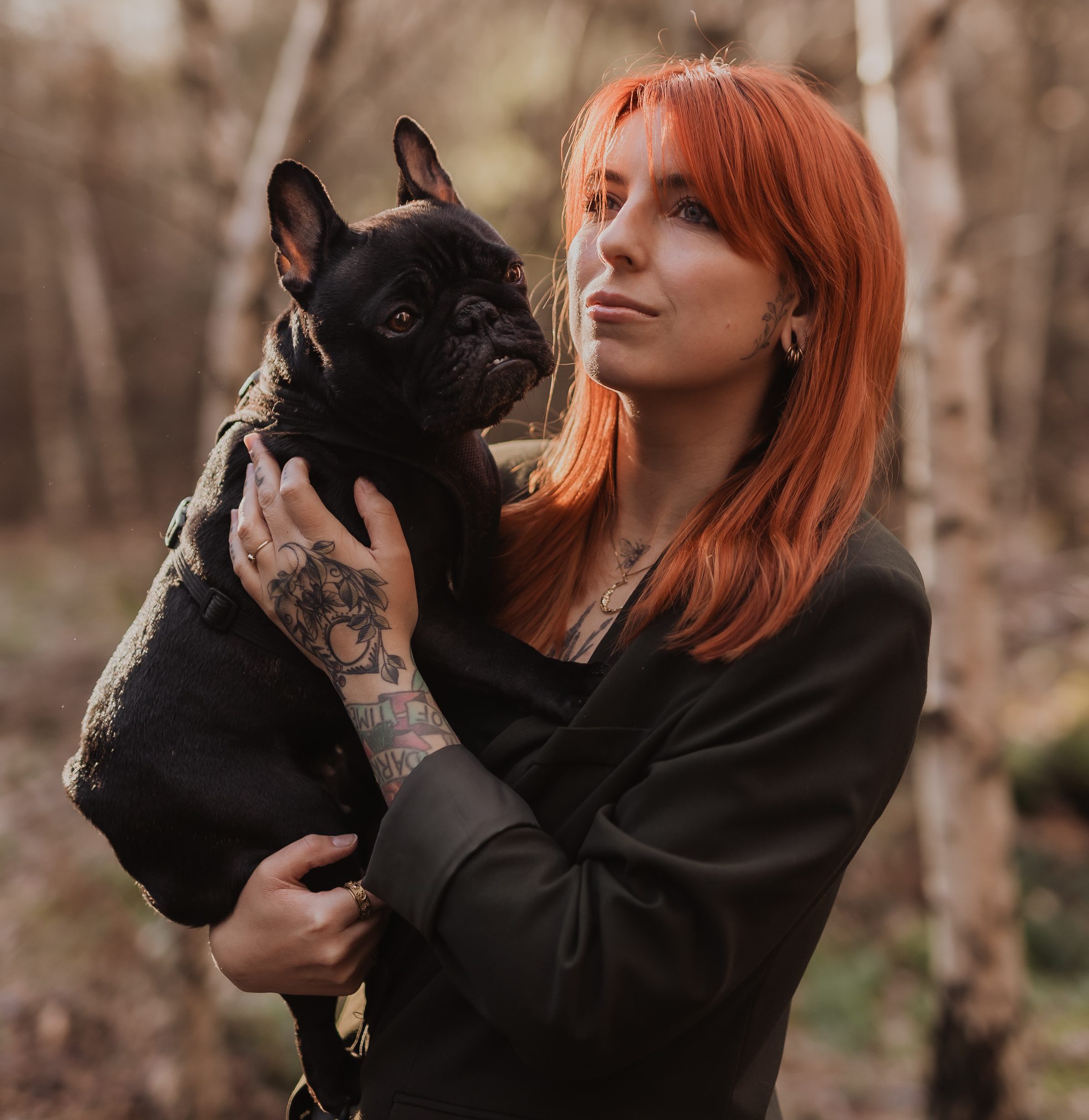 Eine Frau mit langen roten Haaren und Tattoos auf der Hand hält eine schwarze französische Bulldogge. Sie steht in einem Waldstück, trägt eine dunkle Jacke und blickt mit nachdenklichem Gesichtsausdruck zur Seite. Das Sonnenlicht fällt durch die Bäume im Hintergrund.