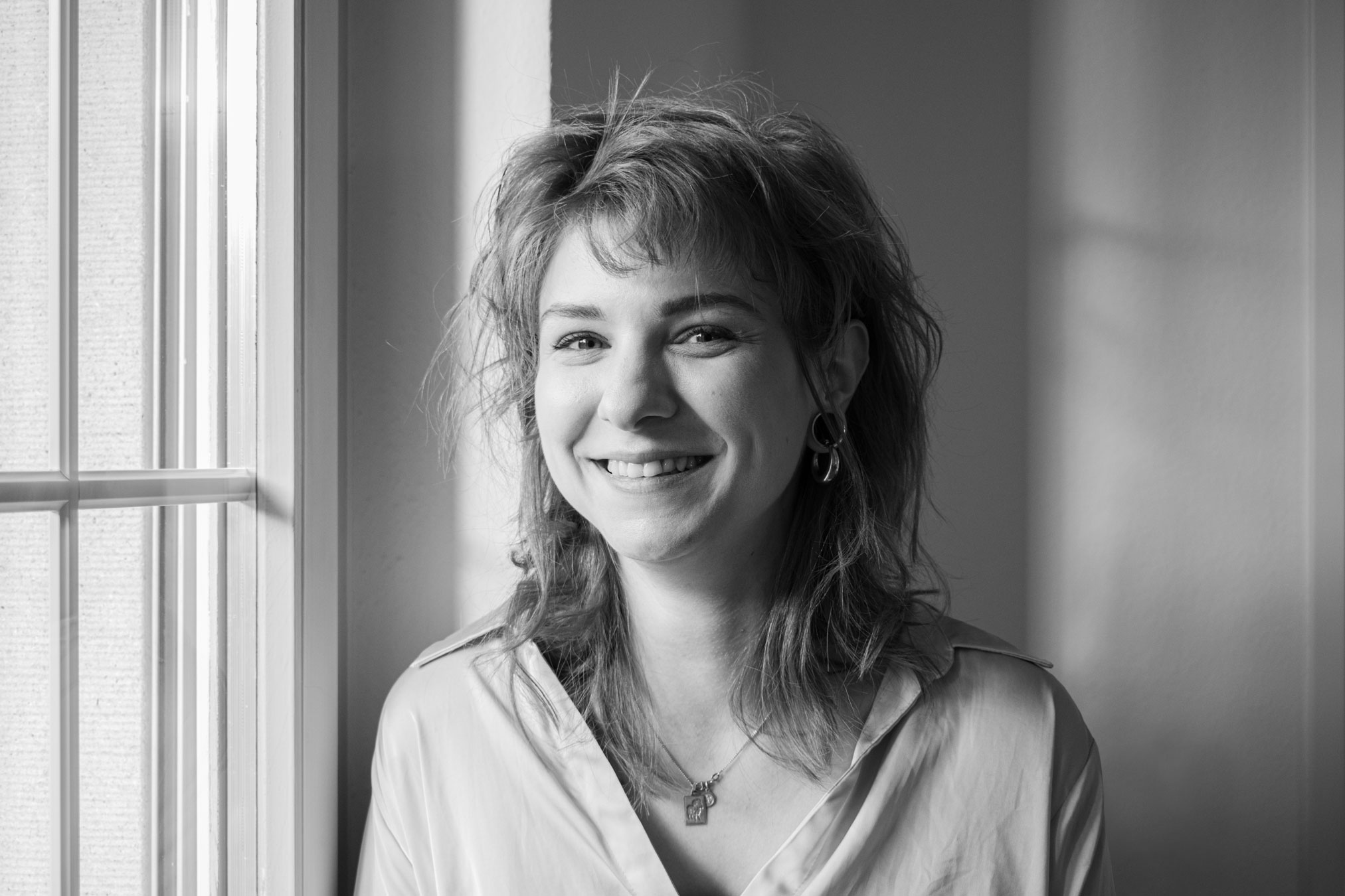 Schwarz-Weiß-Porträt von Tasha Ender mit schulterlangem Haar und einem freundlichen Lächeln. Sie steht neben einem Fenster, dessen Gesicht von natürlichem Licht erhellt wird. Aufgenommen während eines Fotoshootings von einer talentierten Fotografin, trägt sie eine helle Bluse, Ohrringe und eine Halskette. Ihr Haar ist leicht zerzaust.