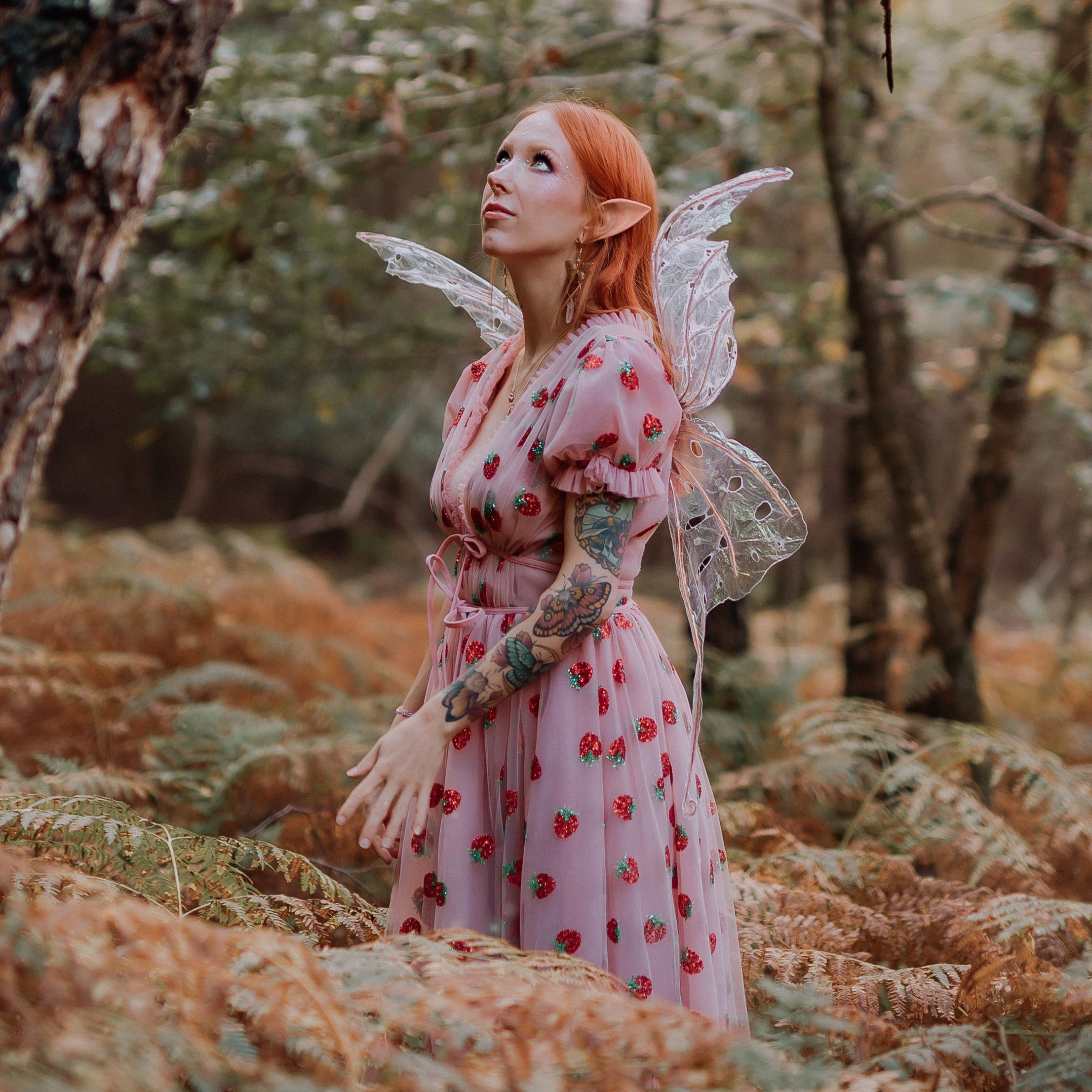 Eine Person mit langen roten Haaren und Elfenohren posiert in einem Wald für eine „Fotoshooting“-Session von Tasha Ender Photography. Sie trägt ein rosa Kleid mit Erdbeermuster und transparente Feenflügel. Ihre bloßen Arme sind mit Tätowierungen verziert und sie blickt nach oben, umgeben von hohen Farnen und Bäumen im Hintergrund.