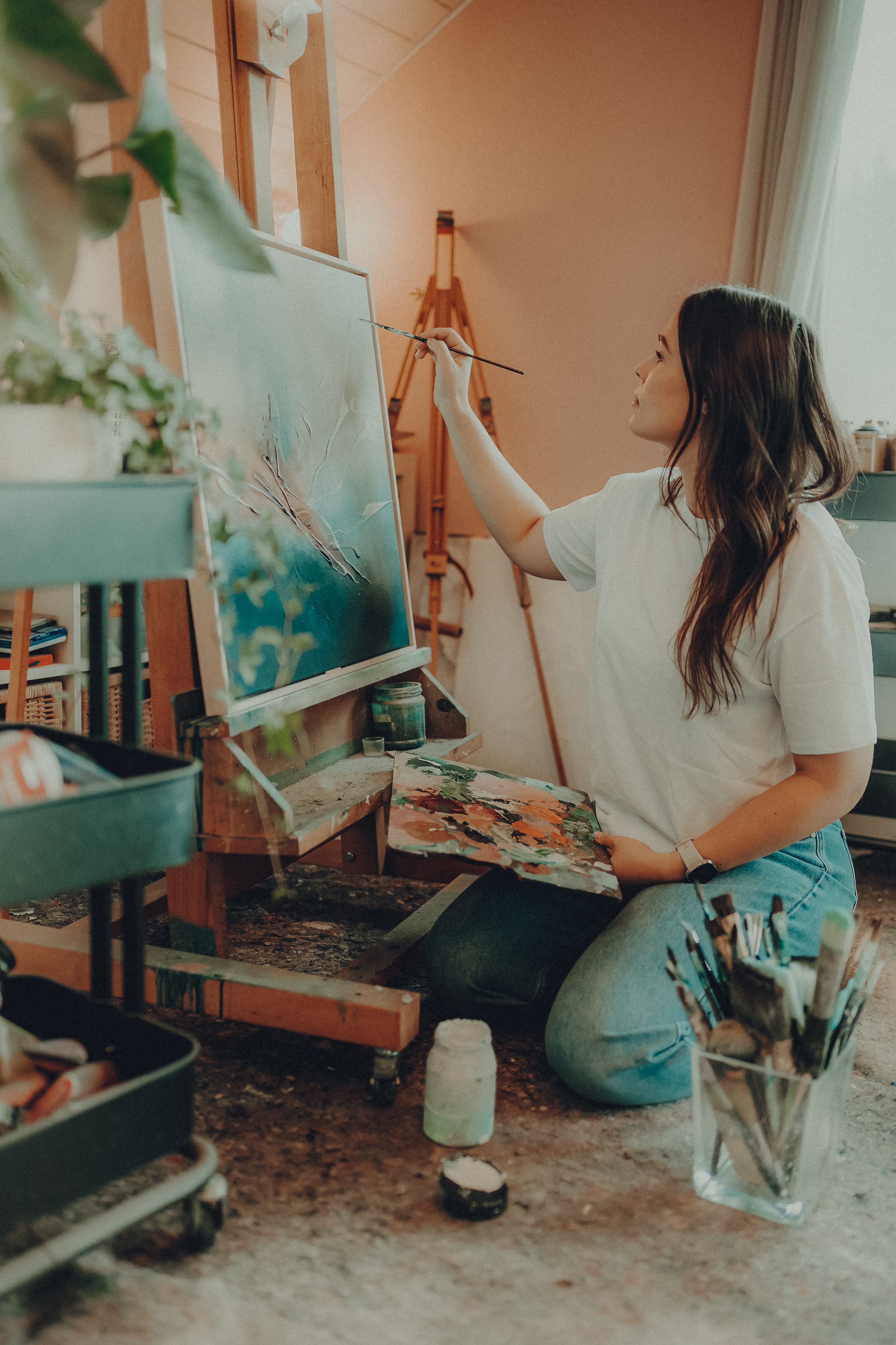 Eine Person mit langen Haaren kniet auf dem Boden und malt auf einer Leinwand, die auf einer Staffelei steht. In der einen Hand hält sie eine Palette mit verschiedenen Farben und in der anderen einen Pinsel. Der für ein Fotoshooting vorbereitete Raum ist mit Malutensilien und Pflanzen gefüllt, was eine kreative und gemütliche Atmosphäre schafft.
