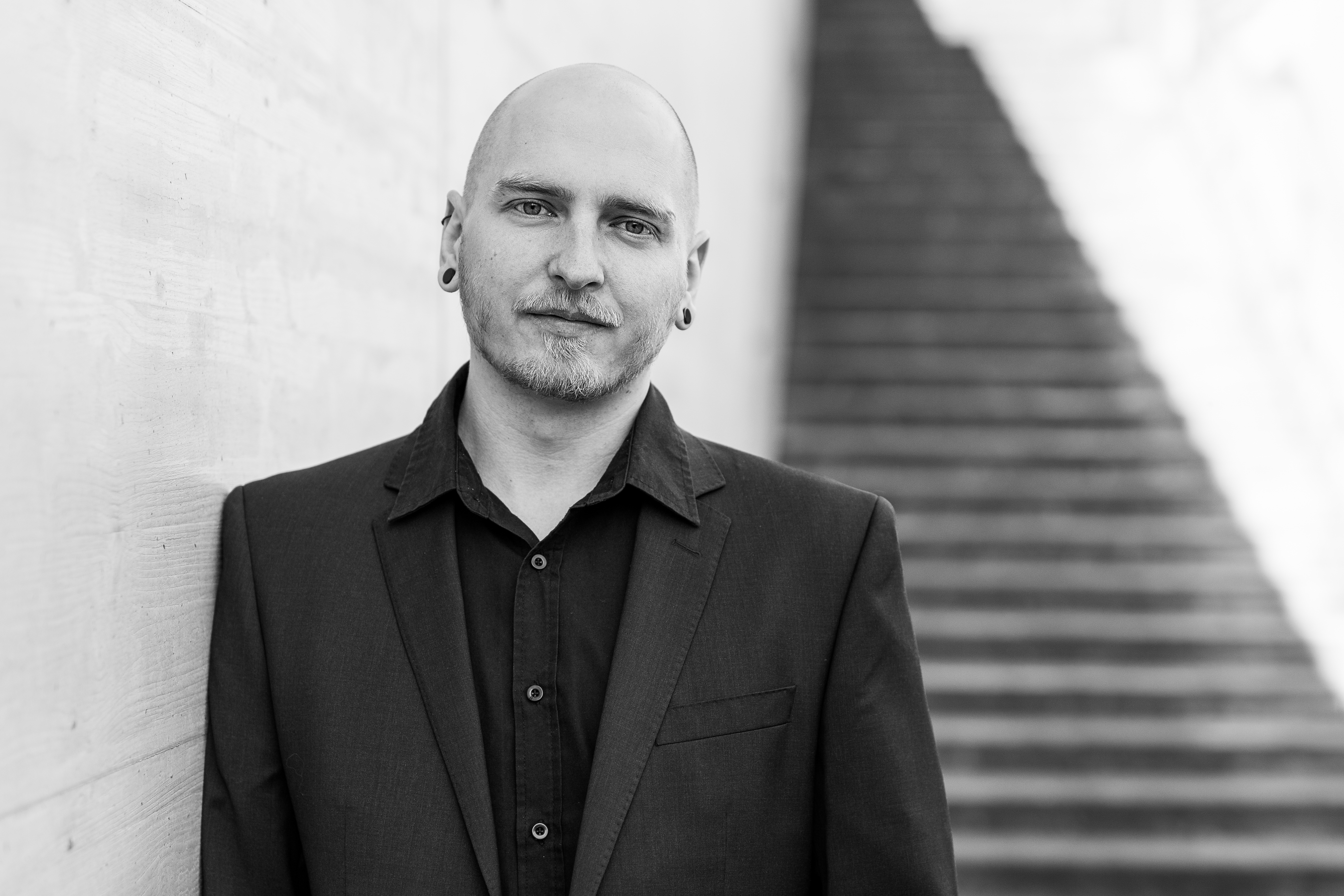 Ein kahlköpfiger Mann mit Ohrlöchern und gestutztem Bart steht in einem dunklen Anzug und Hemd neben einer strukturierten Betonwand. Er blickt während dieser Fotoshooting-Session selbstbewusst in die Kamera. Im Hintergrund ist eine Treppe mit verschwommenen Stufen zu sehen. Schwarz-weißes Porträtfoto, perfekt für seinen Lebenslauf.