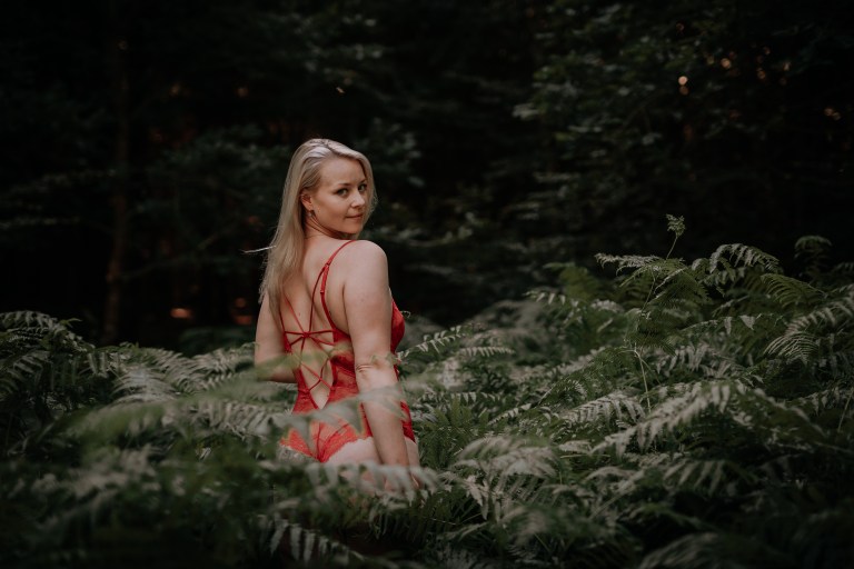 Eine Frau mit langen blonden Haaren steht in einem üppigen Wald, der teilweise von hohen grünen Farnen umgeben ist. Sie trägt ein rotes Trägerkleid und blickt mit einem ruhigen und gelassenen Gesichtsausdruck über ihre Schulter in die Kamera. Der Hintergrund ist mit dichtem, dunklem Laub gefüllt und fängt die Essenz eines sinnlichen Fotoshootings ein.