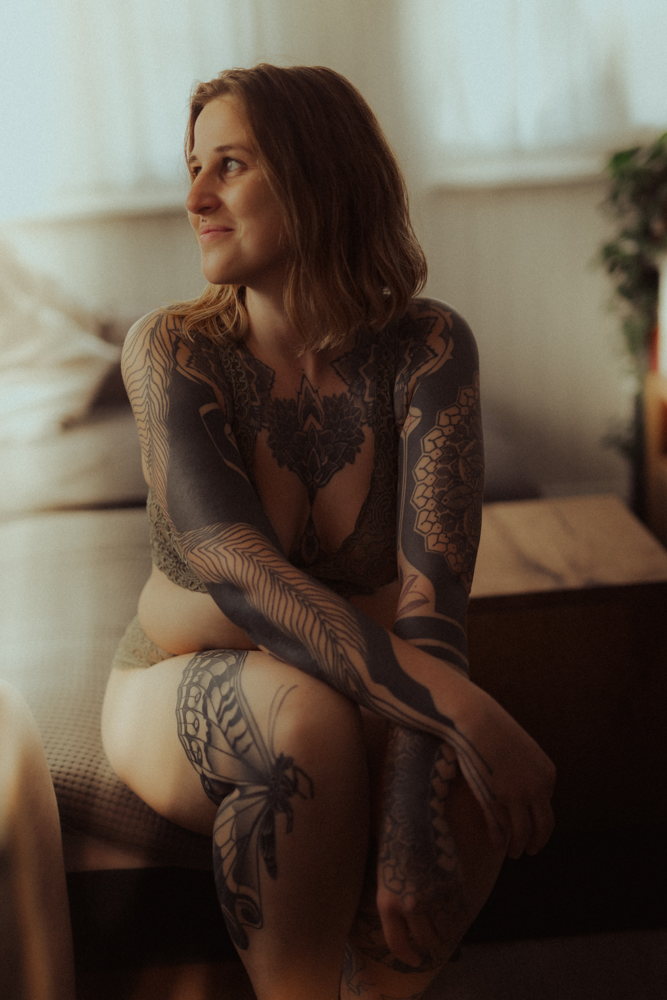 Eine Frau mit langen braunen Haaren und vielen schwarzen Tattoos auf Armen, Brust und Beinen sitzt auf einem Bett in einem Zimmer, das von sanftem, natürlichem Licht erhellt wird. Sie trägt Dessous und blickt nach dem Fotoshooting nachdenklich zur Seite, mit einem leichten Lächeln im Gesicht.