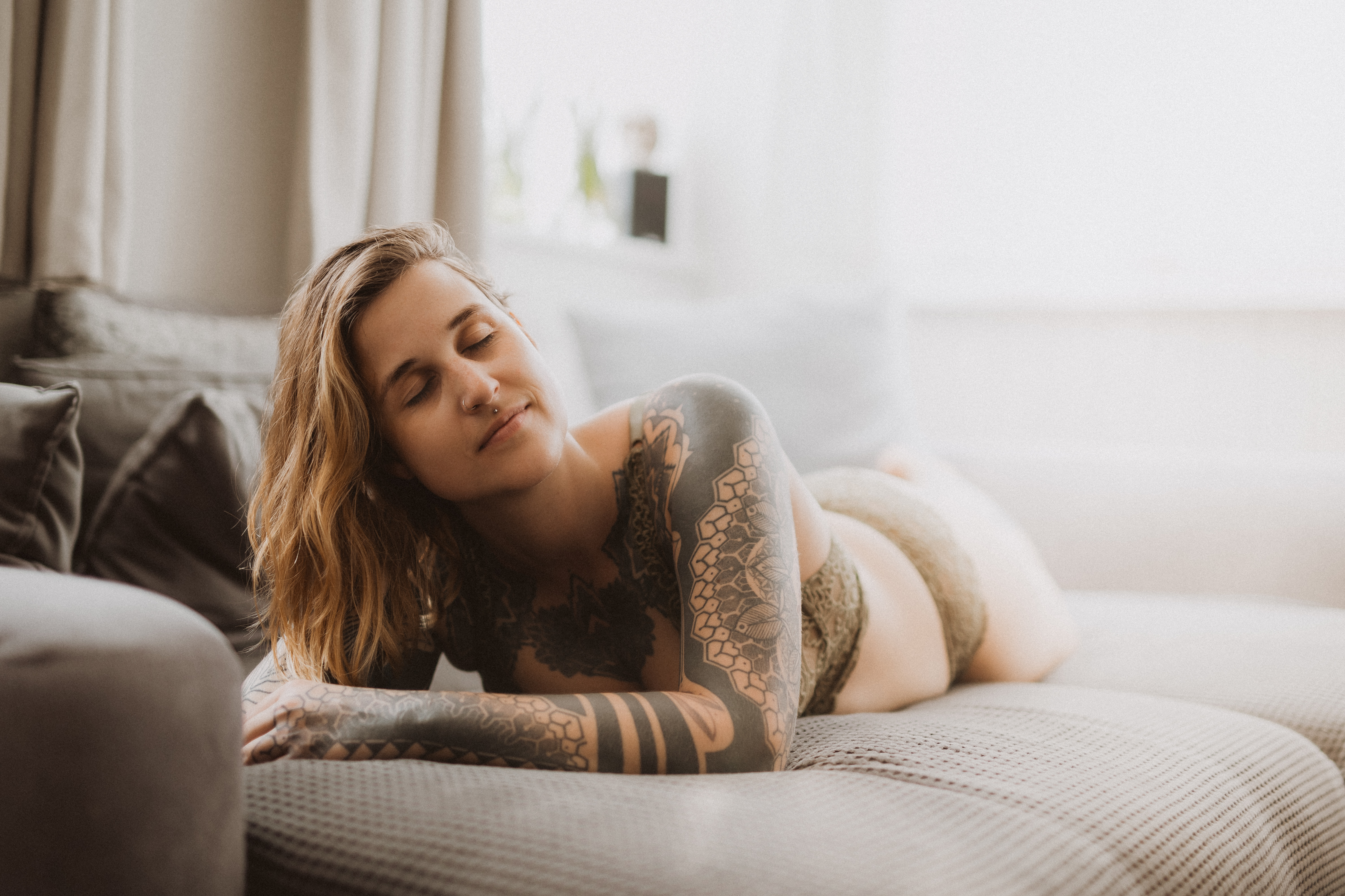 Eine Person mit Tattoos liegt in einem sonnendurchfluteten Raum auf einer grauen Couch und trägt dazu passenden BH und Unterwäsche. Sie hat kurzes, welliges Haar und einen zufriedenen Gesichtsausdruck, die Augen sind geschlossen und ein Arm ruht auf der Couch. Das stilvolle Ambiente dieses Boudoir-Fotoshootings umfasst Vorhänge und Kissen im Hintergrund.