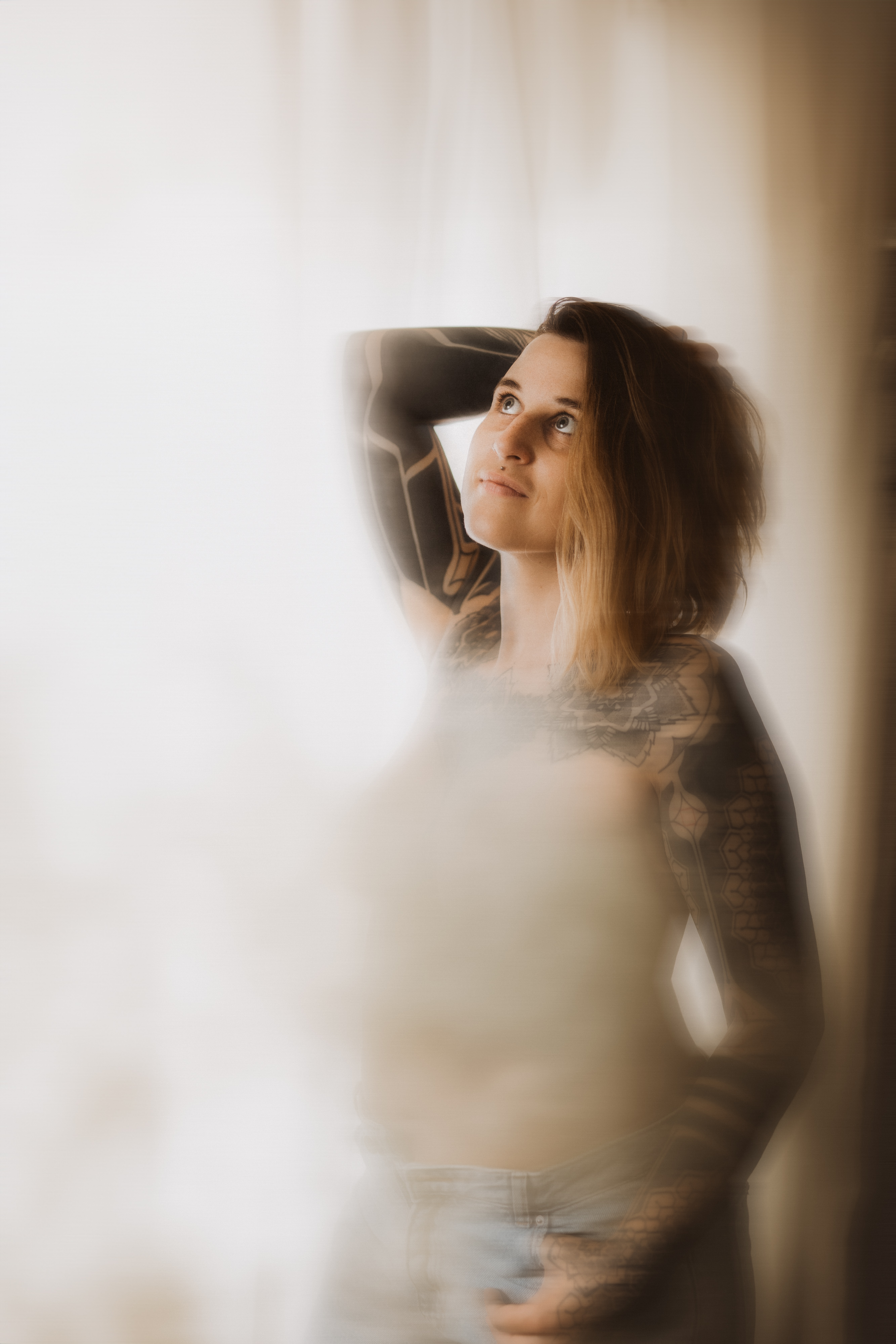 In einer sanft beleuchteten Umgebung steht eine Person mit schulterlangem Haar und Tattoos auf Armen und Brust vor einem ätherischen, verschwommenen Hintergrund. In diesem nachdenklichen Porträt blickt sie nach oben und hat einen Arm so erhoben, dass er ihren Hinterkopf berührt. So fängt sie die Essenz eines natürlichen Moments während des Fotoshootings ein.