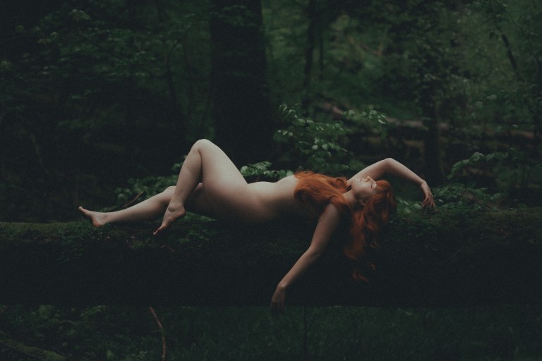 Eine nackte Person mit langen roten Haaren liegt auf einem moosbedeckten Baumstamm in einem dichten, dunklen Wald und erinnert an die Essenz der Natur. Das umgebende Laub ist üppig und grün, wirft tiefe Schatten und schafft eine heitere, natürliche Atmosphäre, die an ein ätherisches Boudoir-Shooting im Freien erinnert.