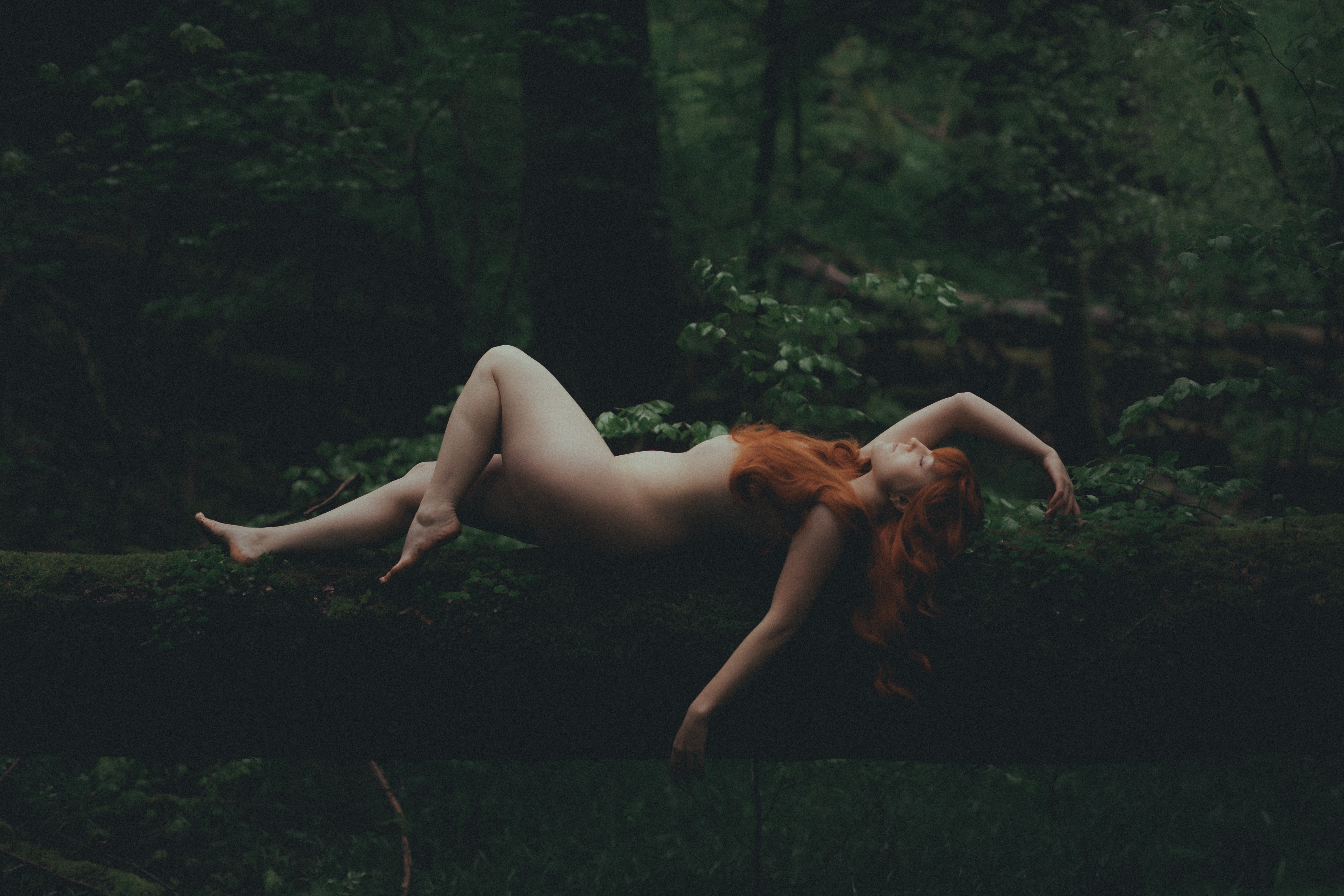 Eine nackte Person mit langen roten Haaren liegt auf einem moosbedeckten Baumstamm in einem dichten, dunklen Wald und erinnert an die Essenz der Natur. Das umgebende Laub ist üppig und grün, wirft tiefe Schatten und schafft eine heitere, natürliche Atmosphäre, die an ein ätherisches Boudoir-Shooting im Freien erinnert.