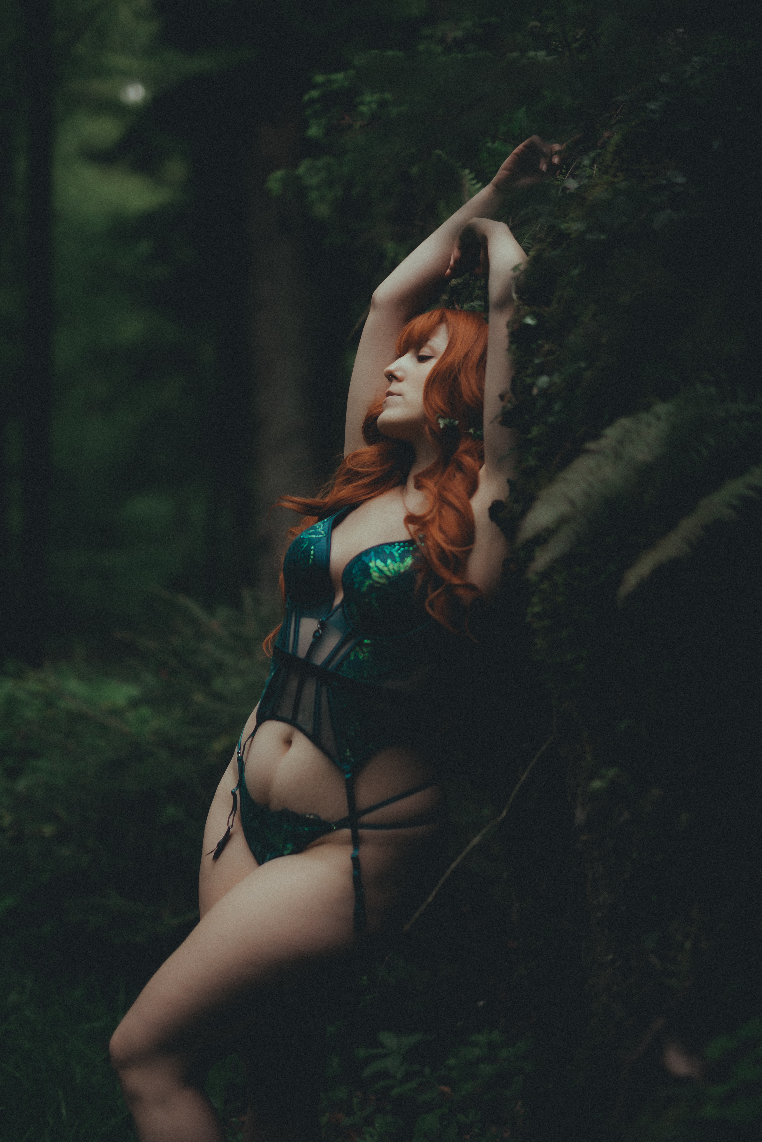 Eine Person mit langen roten Haaren posiert während eines Boudoir-Shootings im Freien in einem Wald und trägt ein grün-schwarzes Dessous-Set. Sie steht mit einem Arm auf dem Kopf und dem anderen über einem moosbedeckten Baum, umgeben von üppigem Grün. Die Beleuchtung ist schwach und stimmungsvoll.