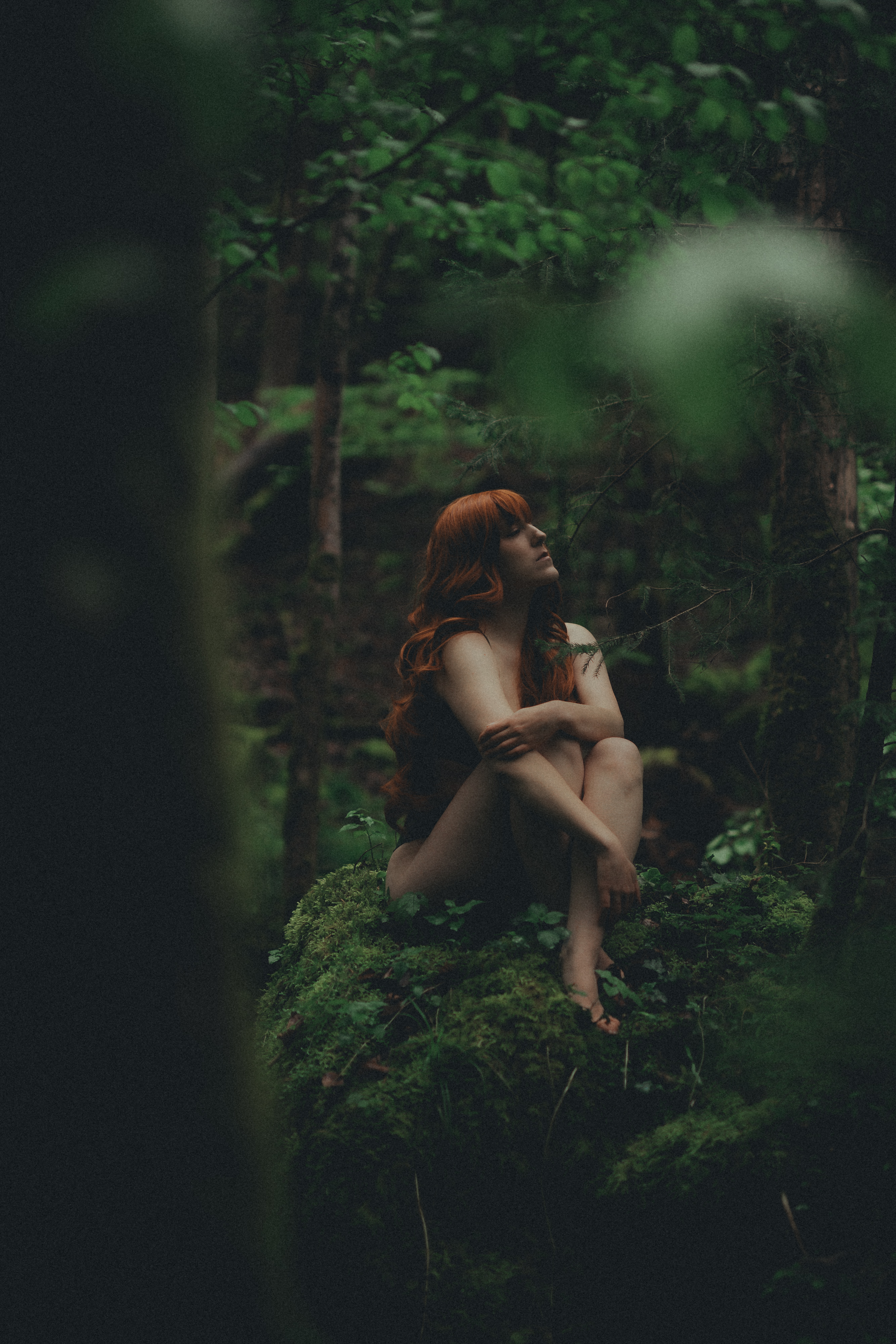 Eine Person mit langen roten Haaren sitzt auf einem moosbedeckten Felsen in einem dichten Wald, als ob sie sich auf ein ruhiges Fotoshooting vorbereitet. Die nackte Figur ist nachdenklich inmitten des üppigen grünen Laubes und der hoch aufragenden Bäume und schafft eine mystische Atmosphäre.