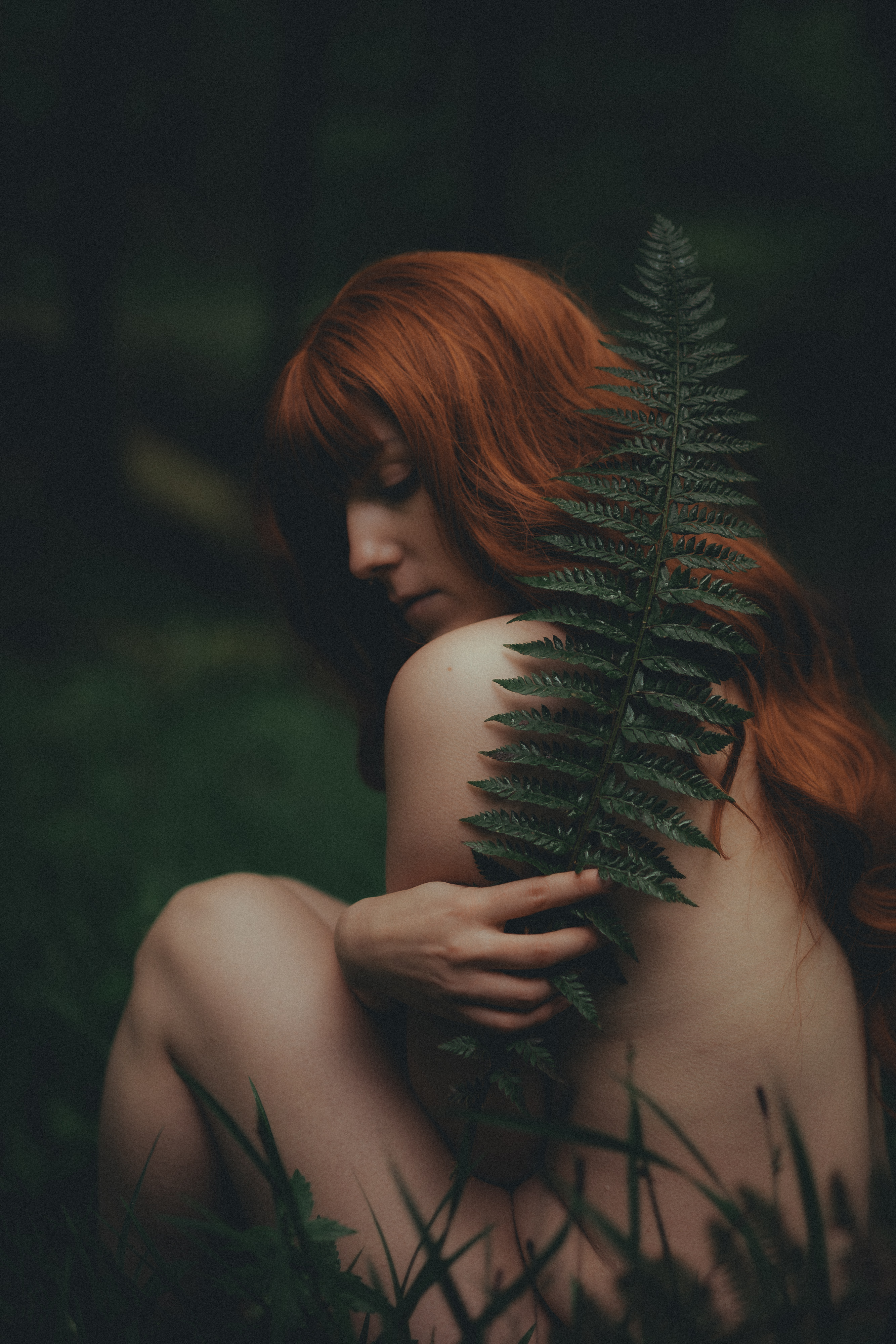 Eine Person mit langen roten Haaren sitzt nackt in einem Wald, teilweise geschützt durch ein großes grünes Farnblatt. Die Szene, aufgenommen während eines künstlerischen Fotoshootings, ist dunkel, mit sanftem natürlichem Licht, das die Person beleuchtet und eine geheimnisvolle und heitere Atmosphäre schafft.