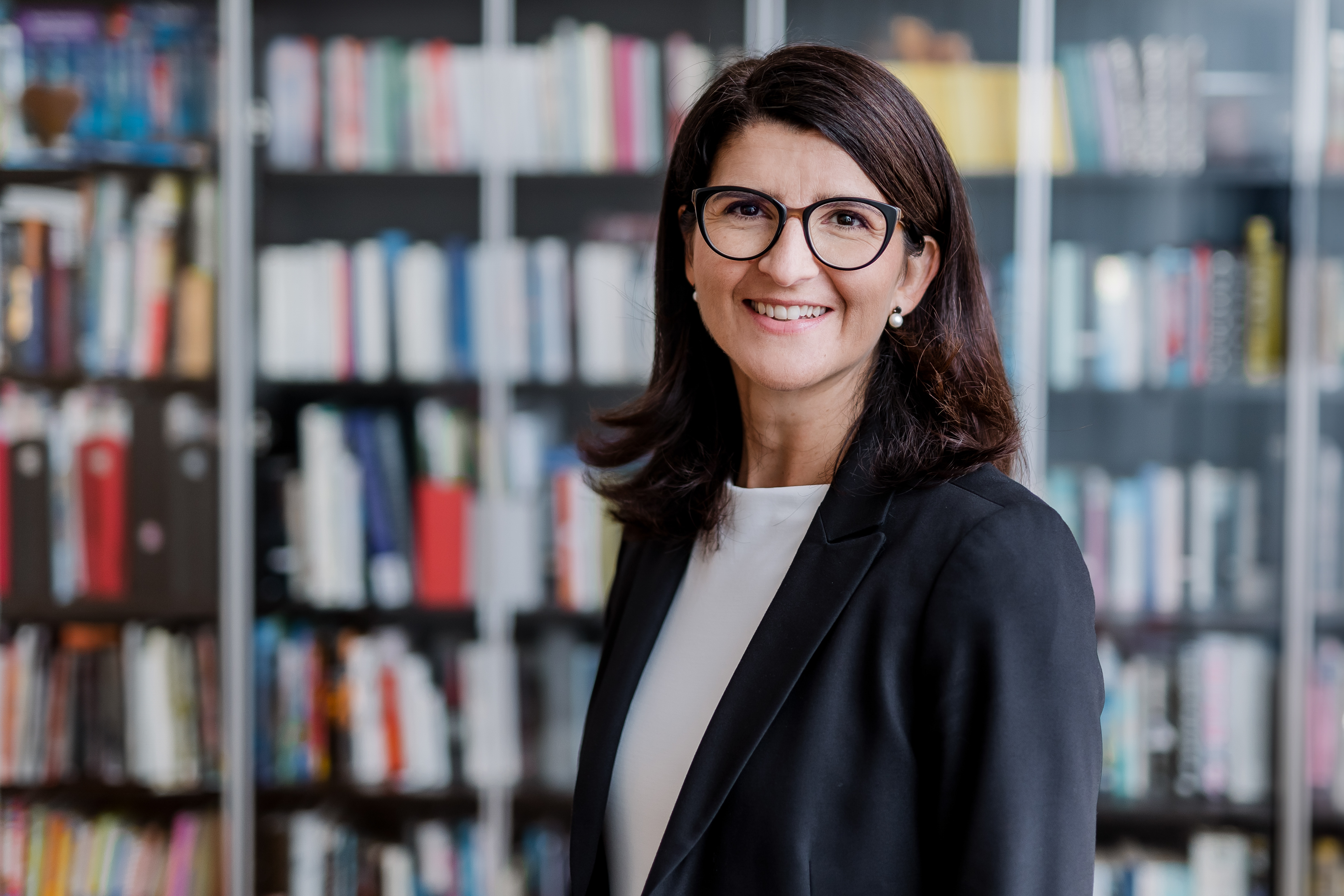 Eine Frau mit schulterlangem braunem Haar und Brille steht lächelnd vor einem Bücherregal voller Bücher. Sie trägt einen schwarzen Blazer über einem weißen Oberteil. Der Hintergrund ist leicht unscharf und hebt sie als Hauptmotiv hervor – ein ideales Bewerbungsbild für jeden Lebenslauf.