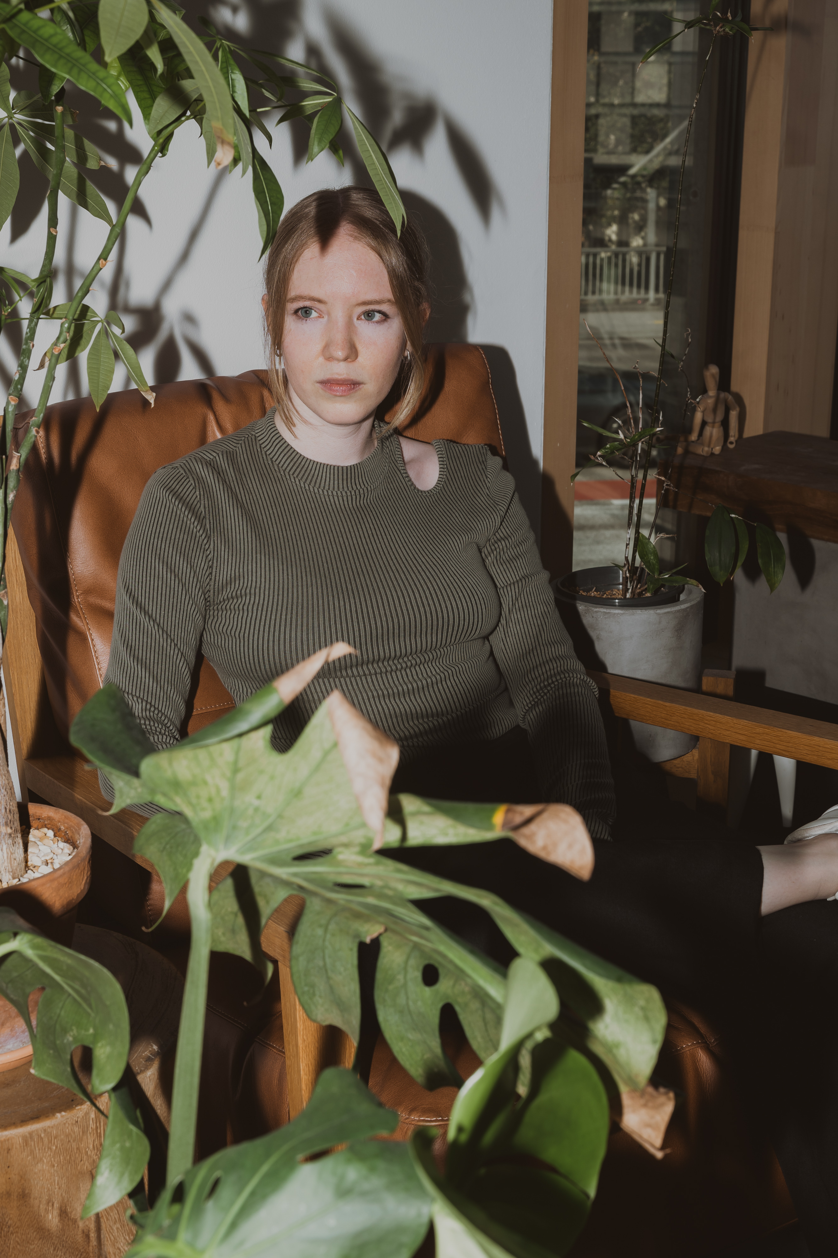 Eine Frau mit rotbraunem Haar sitzt in einem braunen Ledersessel und trägt einen dunkelgrünen Rippenpullover mit Cut-Out-Detail. Umgeben von verschiedenen Topfpflanzen und in Sonnenlicht getaucht, das durch ein nahes Fenster fällt, posiert sie für ihr Bewerbungsbild. Ihr Gesichtsausdruck ist ruhig und nachdenklich.