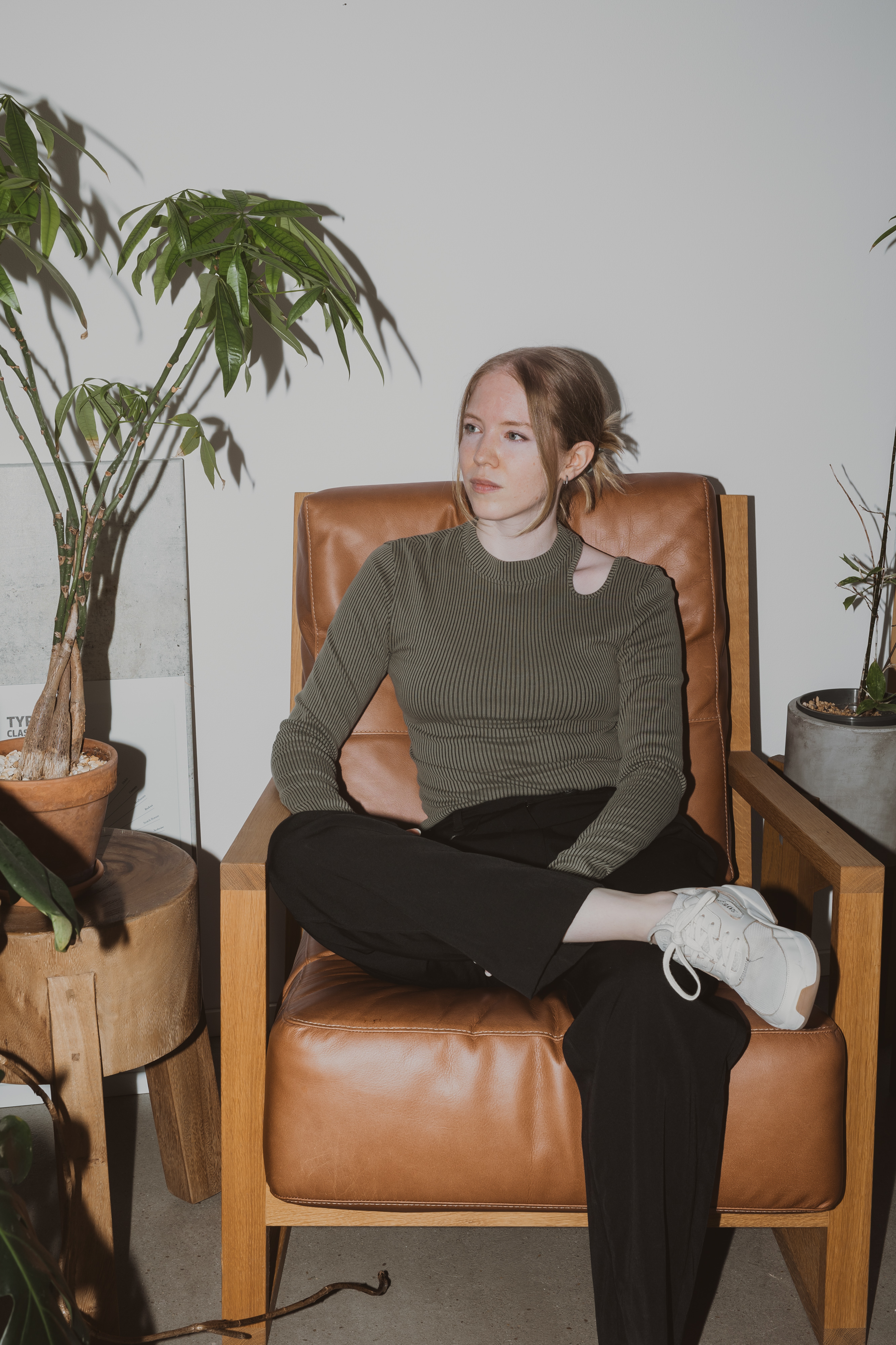 Eine Frau mit heller Haut und schulterlangem Haar sitzt auf einem hellbraunen Lederstuhl und blickt in einem ruhigen Porträt nach links. Sie trägt einen grünen Rippenpullover und eine schwarze Hose und hat ihr rechtes Bein über das linke geschlagen. Um sie herum stehen Topfpflanzen auf einem Holzhocker und dem Boden, die eine ruhige, baden-ähnliche Atmosphäre schaffen.