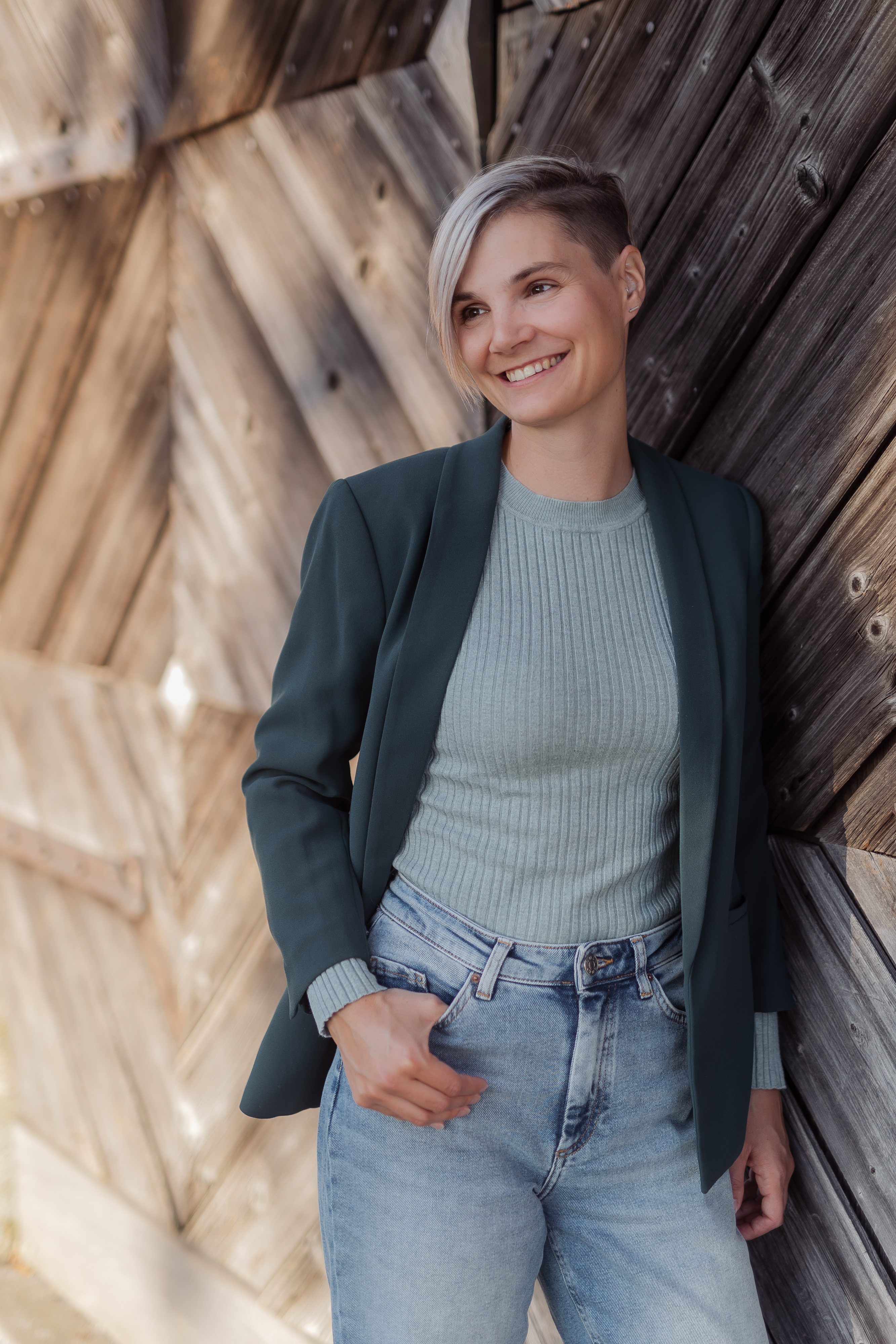 Eine Person mit kurzen, hellen Haaren lehnt lächelnd an einer Holzwand. Sie trägt einen grünen Blazer über einem hellen Rippenpullover und Jeans und ist damit ein ideales Bewerbungsbild. Das Sonnenlicht schafft eine warme und entspannte Atmosphäre, perfekt für ein Bewerbungsfoto.