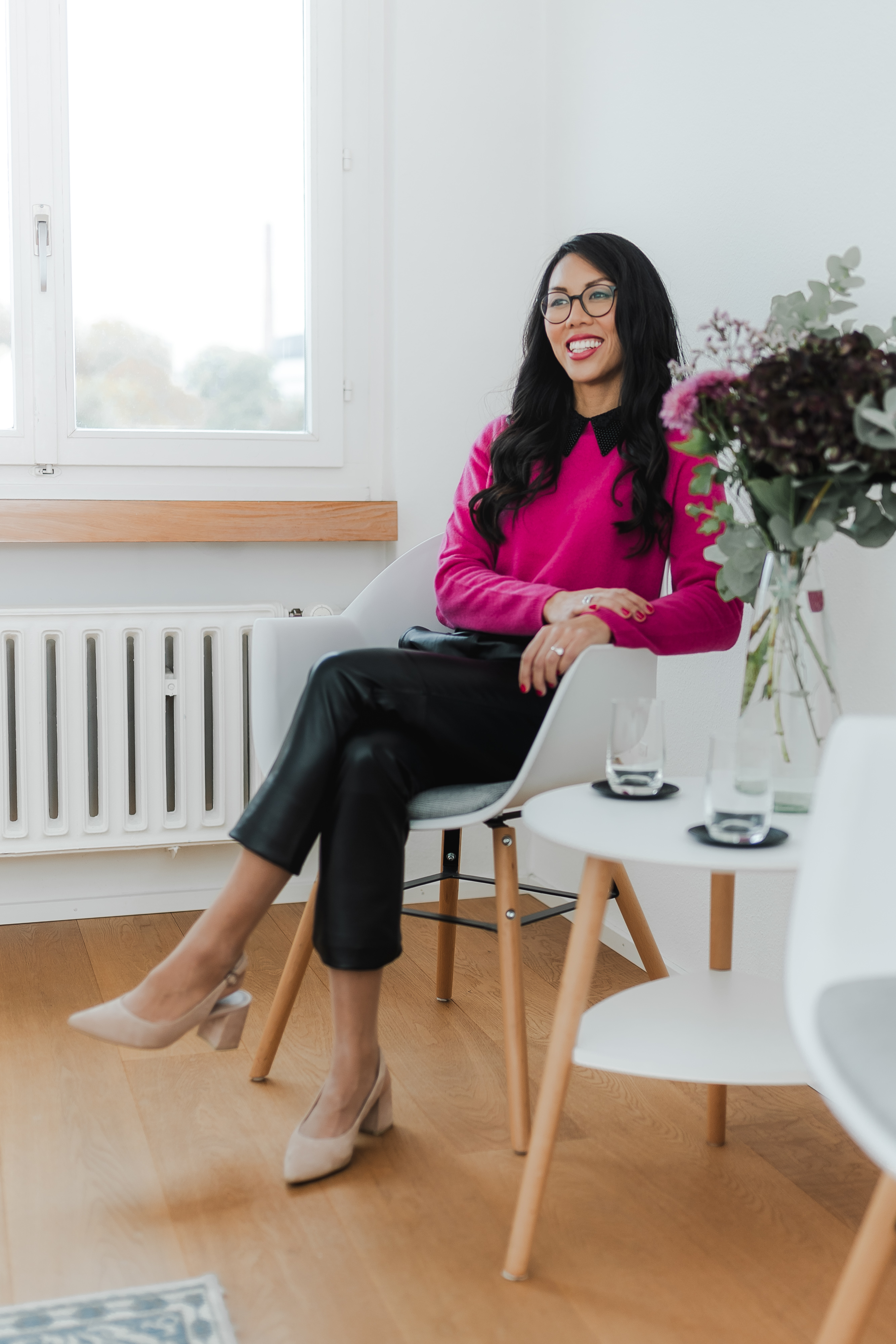 Eine Frau mit langen dunklen Haaren und Brille sitzt gemütlich auf einem modernen weißen Stuhl für ihr Bewerbungsbild. Sie trägt einen leuchtend rosa Pullover, schwarze Lederhosen und beige Pumps. Neben ihr steht ein kleiner Tisch mit Wassergläsern und einer Blumenvase. Der Raum ist in hellen Farben eingerichtet.
