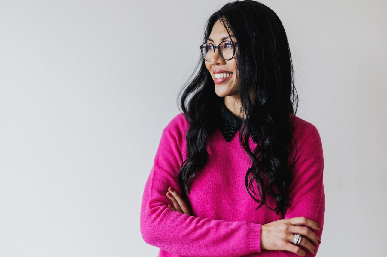 Fotografin Tasha Ender, mit langen dunklen Haaren und Brille, lächelt und schaut nach links. Sie trägt einen leuchtend rosa Pullover und hat die Arme vor einer schlichten weißen Wand verschränkt, wahrscheinlich aufgenommen während eines Fotoshootings.