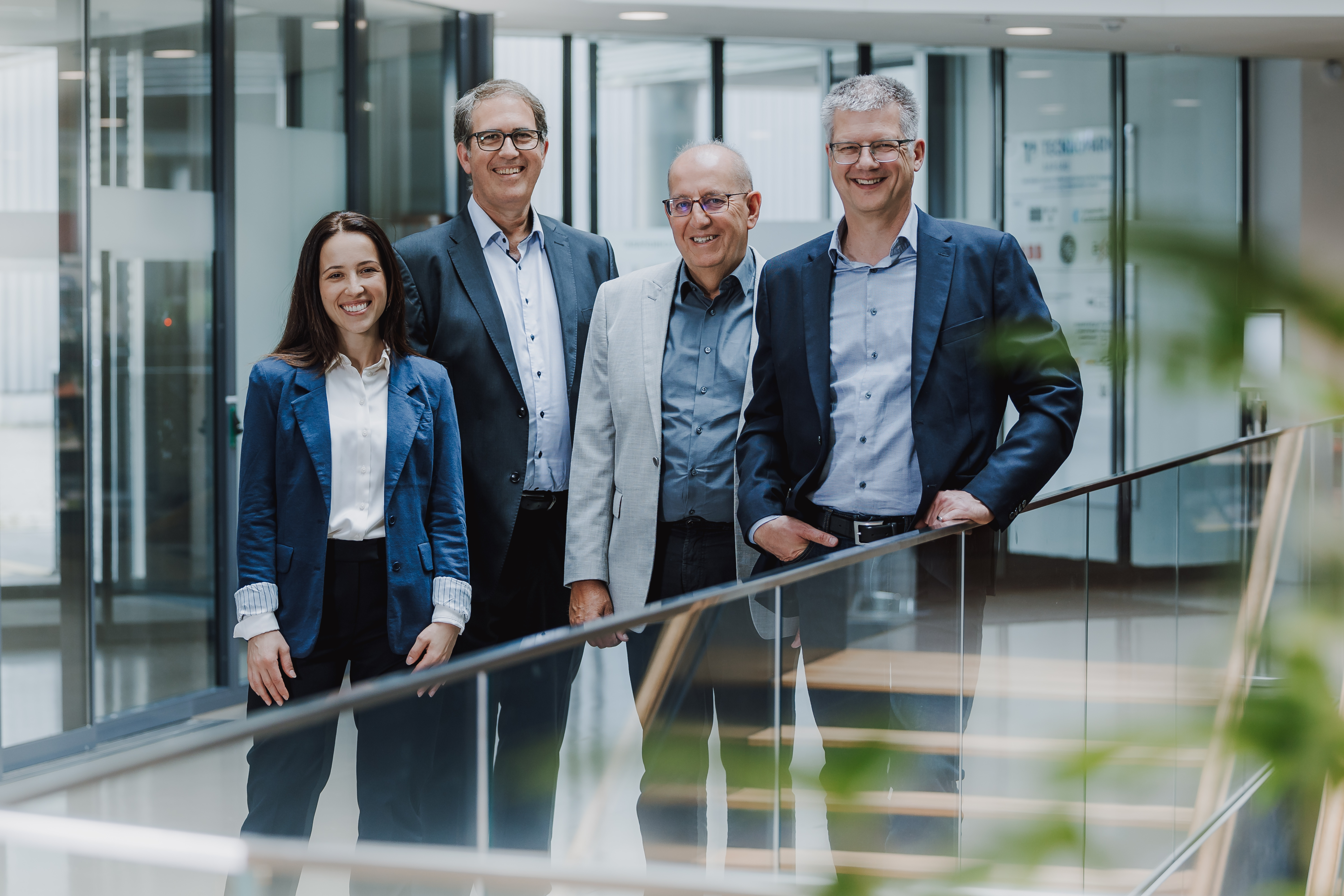 Eine Gruppe von vier Personen, drei Männer und eine Frau, stehen zusammen in einem modernen Bürogebäude. Sie tragen alle Business-Kleidung und lächeln in die Kamera, als ob sie sich auf ein Bewerbungsfoto-Shooting vorbereiten würden. Im Hintergrund sind Glaswände und ein Geländer zu sehen.