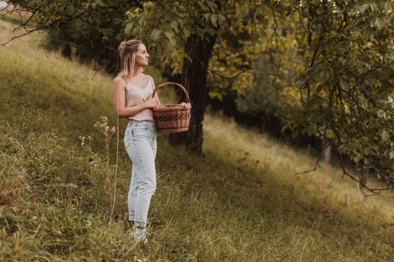 Eine Frau steht auf einem grasbewachsenen Hügel unter einem Baum und hält einen Weidenkorb. Sie trägt ein helles Tanktop und Jeans und blickt nachdenklich in die Ferne. Der Hintergrund besteht aus grünem Laub und einer sanft abfallenden Landschaft, perfekt für Portraitaufnahmen oder Content-Fotografie für Website-Fotos.