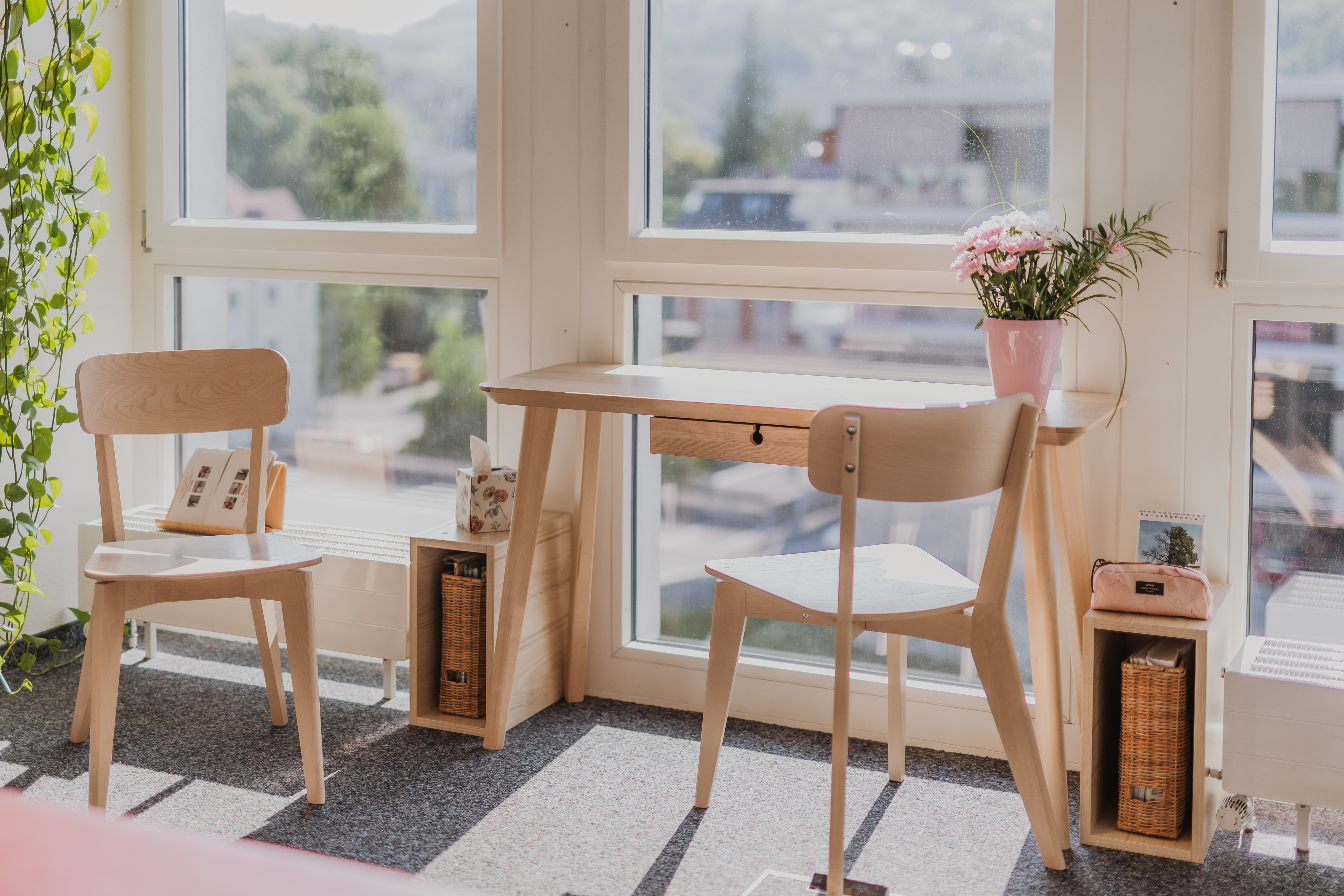 Ein heller Raum mit großen Fenstern und Blick auf die Stadt, perfekt für Firmenfotografie. Drinnen steht ein minimalistischer Holzschreibtisch mit zwei Stühlen. Auf einem Stuhl liegt ein Kissen mit Katzenmotiv. Auf dem Schreibtisch steht eine Vase mit rosa Blumen und darunter Weidenkörbe.