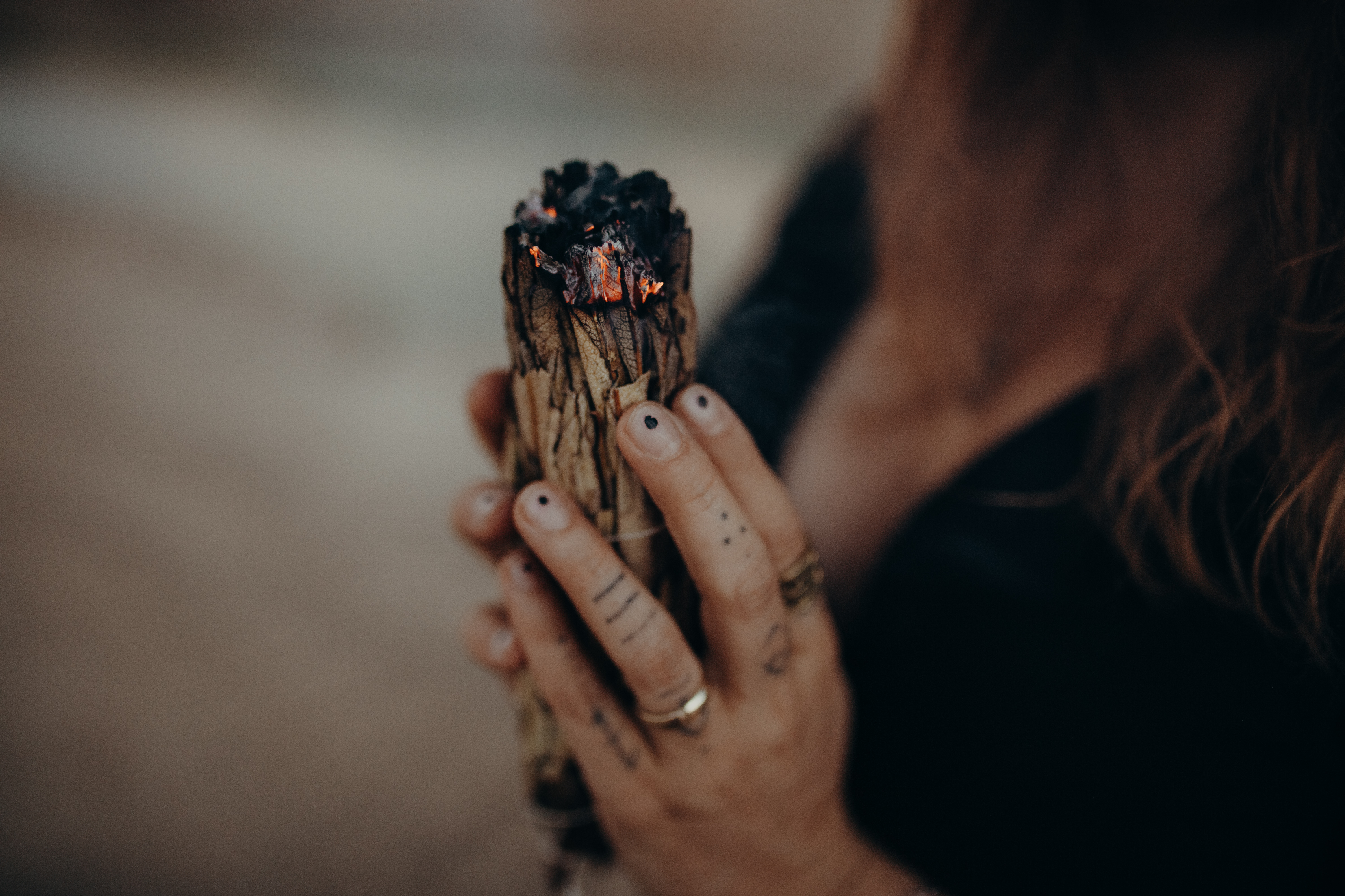 Eine Person mit Tattoos an den Fingern und Ringen an der Hand hält ein brennendes Bündel Salbei. Das Hochzeitsfotografie-Bündel von Tasha Ender Photography stößt kleine Glut und Rauch aus. Der Hintergrund ist unscharf und das Gesicht der Person ist auf dem Bild nicht zu sehen.