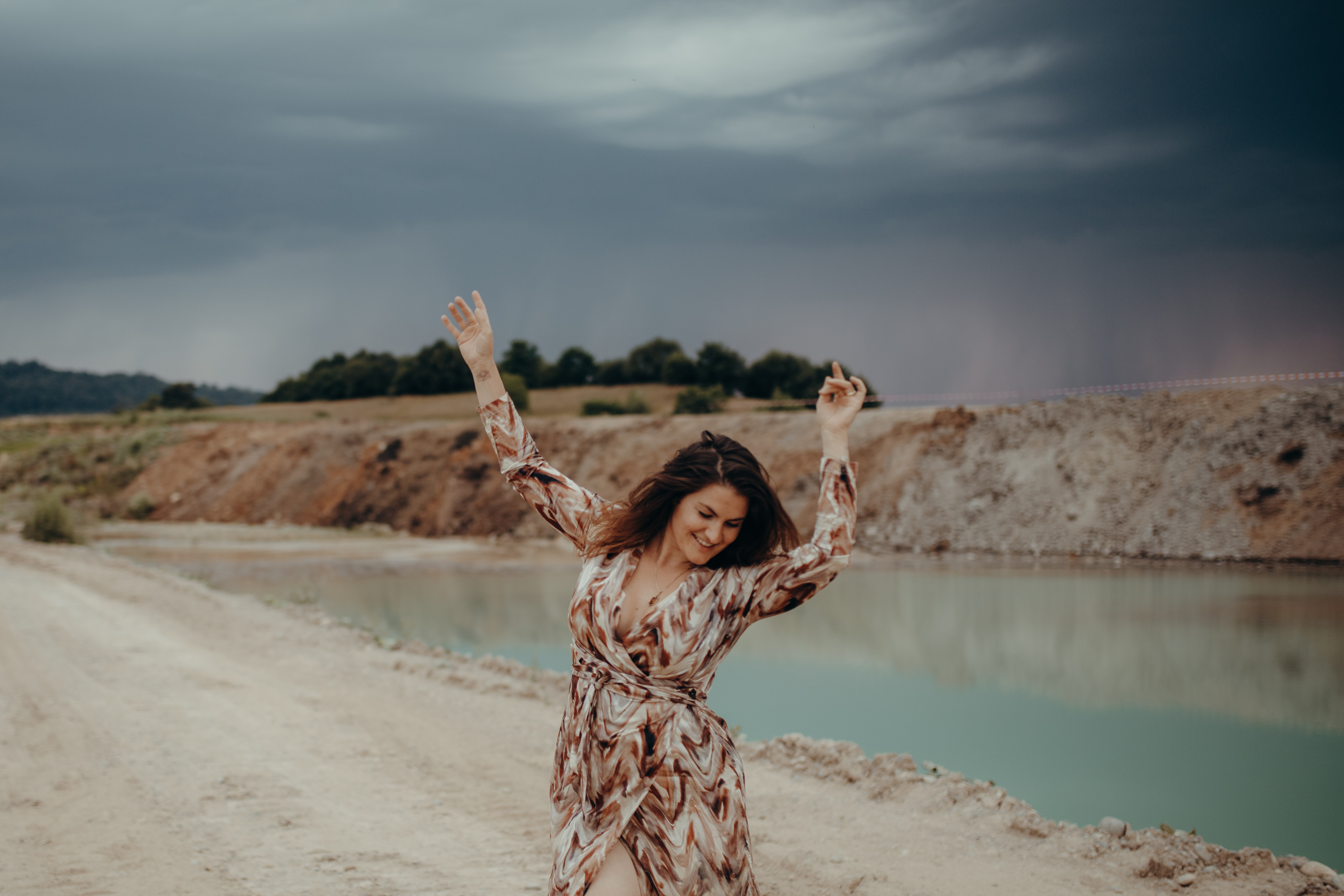 Eine Frau steht auf einem Feldweg neben einem Gewässer im Aargau, im Hintergrund ziehen dunkle Gewitterwolken auf. Sie trägt ein gemustertes, langärmeliges Kleid, hat die Arme freudig in die Höhe gehoben und lächelt – ein perfektes Porträt, aufgenommen von einem erfahrenen Fotografen.