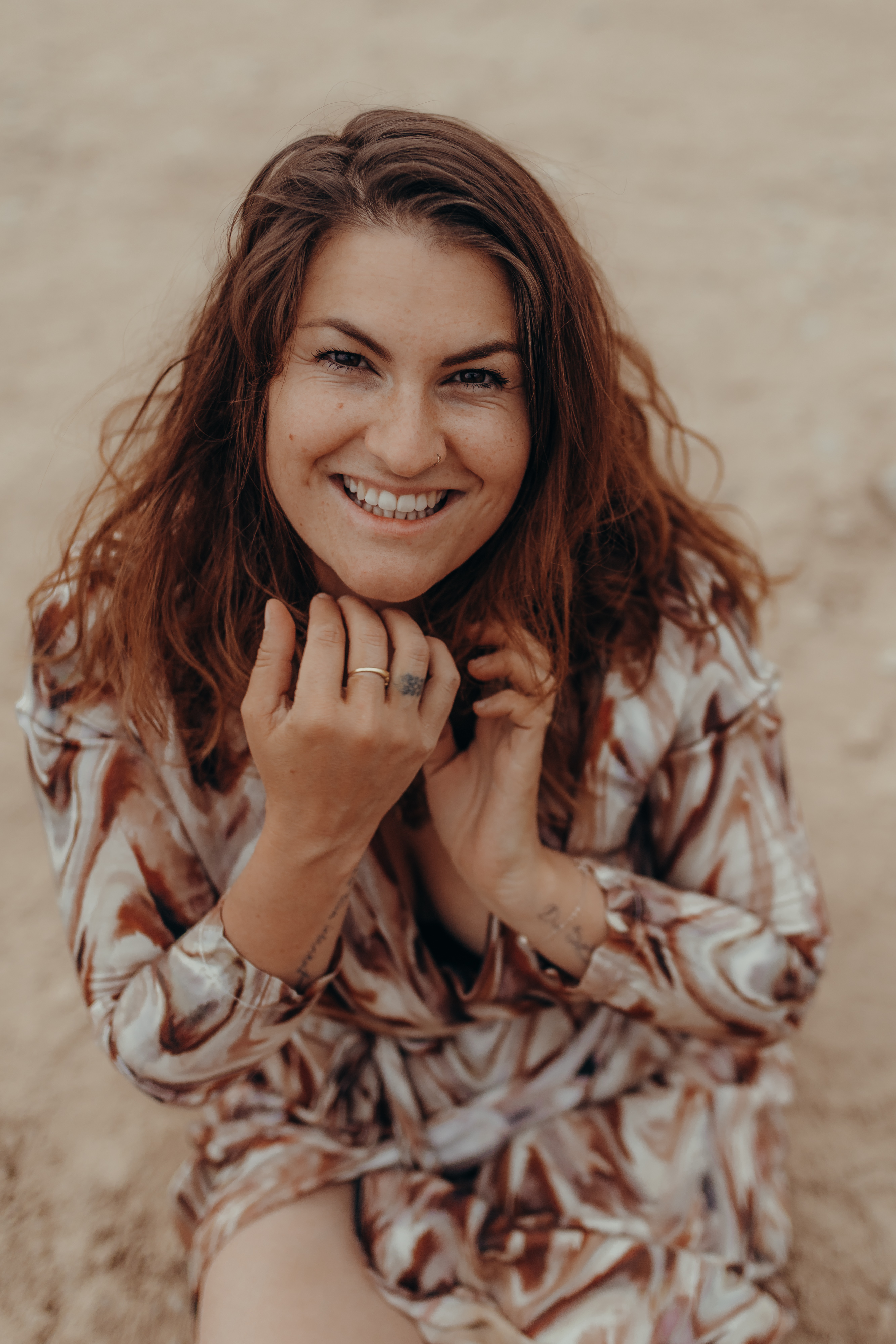 Eine Frau mit welligem, schulterlangem braunem Haar lächelt während eines Fotoshootings in die Kamera. Sie trägt ein gemustertes Kleid und hält ihre Hände vor ihr Gesicht, so dass ein Ring an ihrem Finger zu sehen ist. Der Hintergrund ist eine sandige Außenfläche, die der Fotograf perfekt eingefangen hat.