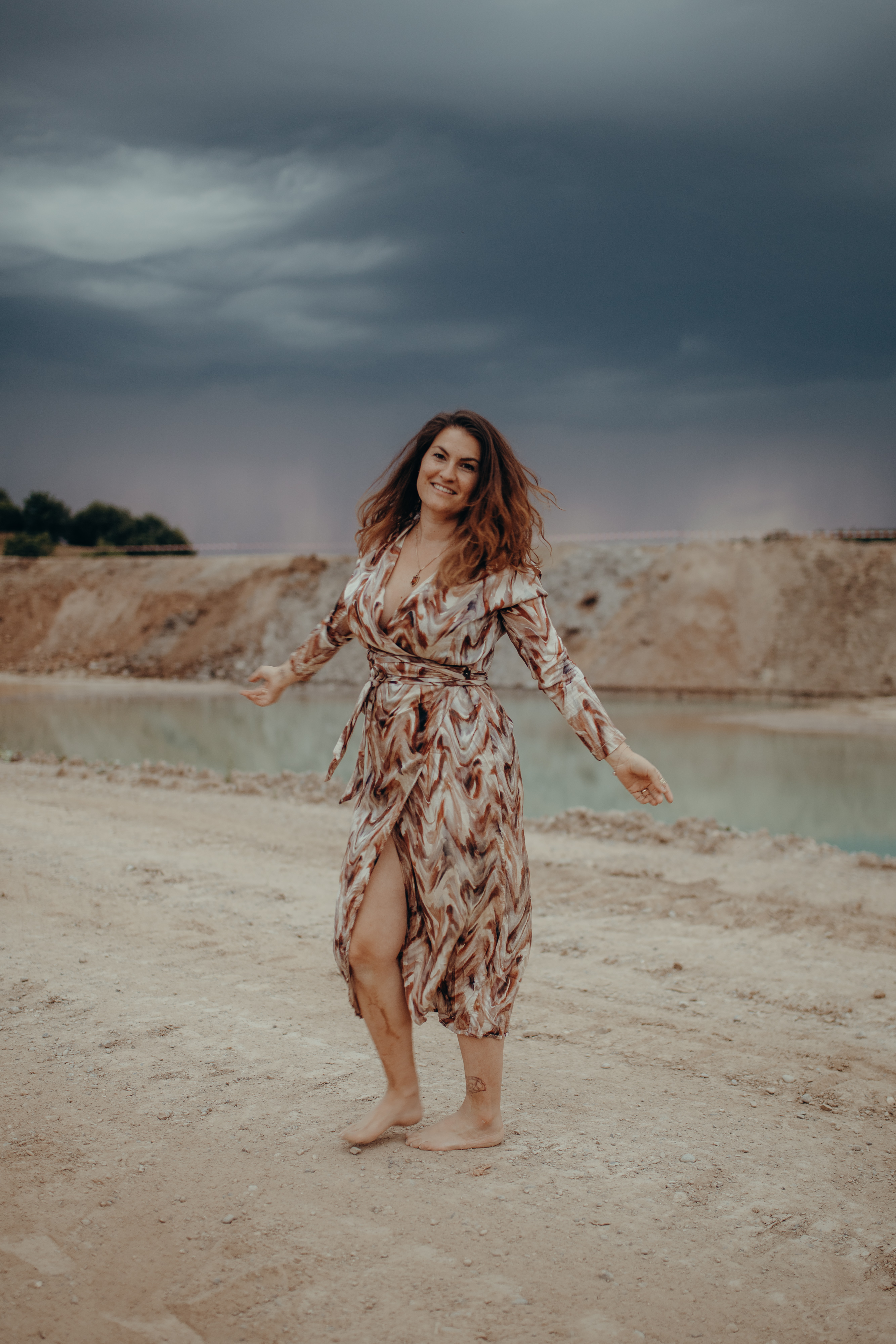 Eine Person mit mittellangem Haar und lächelndem Gesichtsausdruck steht barfuss auf einer felsigen Fläche in der Nähe eines Gewässers. Sie trägt ein gemustertes Kleid mit Gürtel in der Taille. Der Himmel hinter ihr ist mit dunklen Wolken bedeckt, was auf einen bevorstehenden Sturm hindeutet. Dieses eindrucksvolle Porträt fängt die Essenz des Inhalts ein. Fotografie von Aargaus landschaftlicher Schönheit.