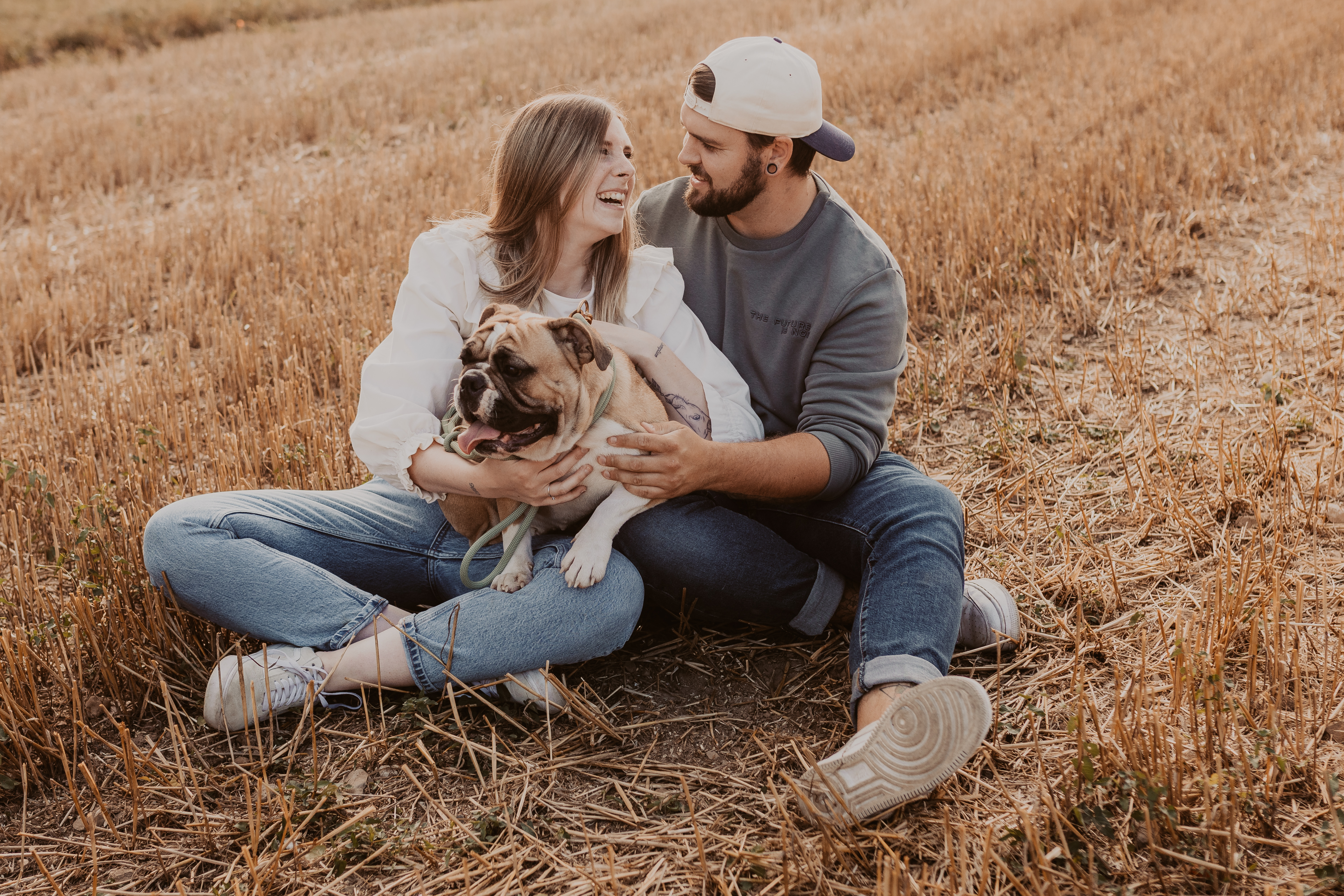 Ein Paar sitzt auf einer Wiese und lacht ausgelassen. Auf dem Schoss der Frau sitzt eine Bulldogge. eS herrscht eine warme und lockere Stimmung während des Fotoshootings mit dem Paar samt Hund.