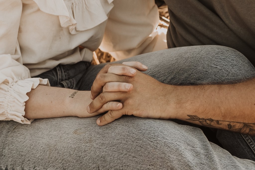Zwei Personen, eine in einem weißen, gerüschten Langarmshirt und die andere in einem grauen Shirt und Jeans, halten sich bei einem Paarshooting an den Händen. Die Person in Weiß hat ein kleines Tattoo am Handgelenk, während die Person in Grau ein Tattoo auf dem Unterarm hat – perfekt eingefangen von der Fotografin.