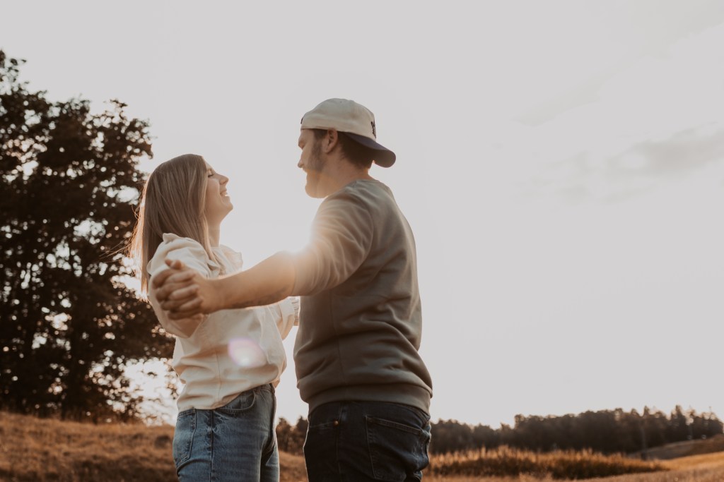 Ein Paar steht bei Sonnenuntergang auf einem offenen Feld, hält sich an den Händen und lächelt sich an. Die Frau trägt ein weißes Oberteil und Jeans, während der Mann ein Langarmshirt, Jeans und eine verkehrt herum aufgesetzte Baseballkappe trägt. Im Hintergrund sind Bäume zu sehen, die den perfekten Moment während ihres Fotoshootings mit einer erfahrenen Fotografin einfangen.