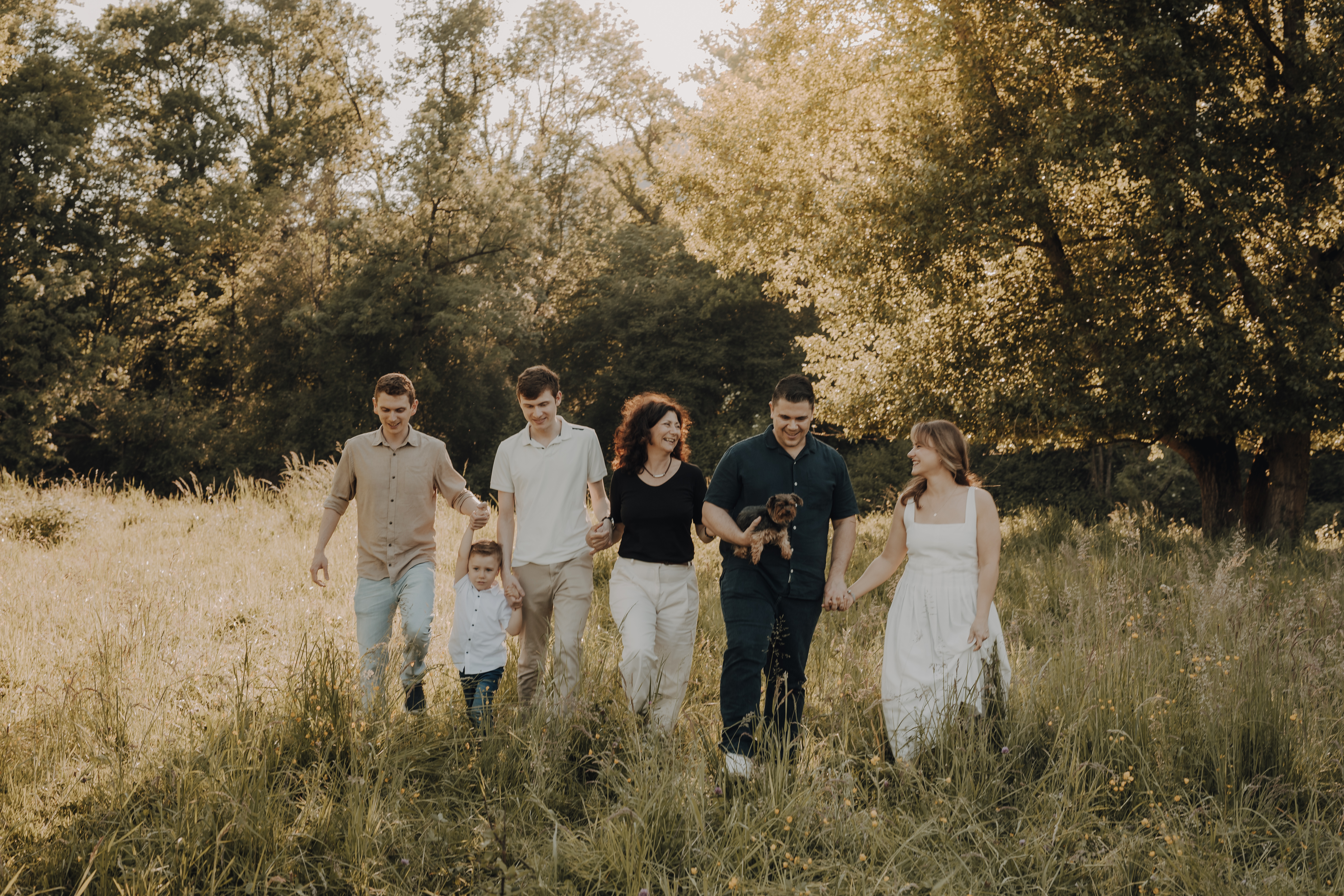 Eine Gruppe von sechs Personen, darunter ein Kind und eine Person mit einem kleinen Hund, gehen durch eine sonnenbeschienene Wiese, die von hohem Gras und Bäumen umgeben ist. Sie lächeln alle, halten sich an den Händen und genießen gemeinsam die Natur an einem hellen Tag. Es sieht aus wie die perfekte Szene für Familienfotos mit einer Fotografin in Baden.