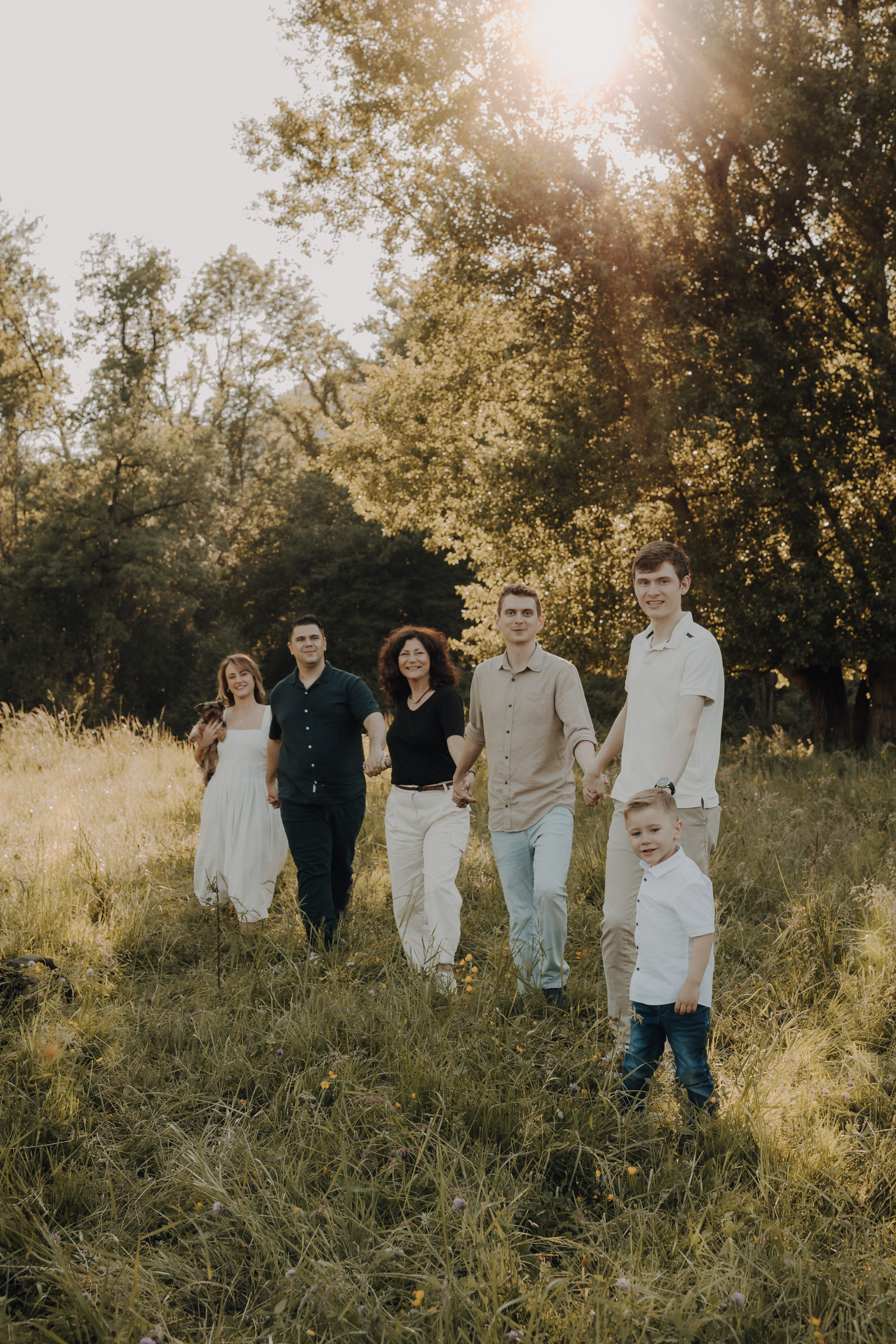 Eine Gruppe von sechs Personen, darunter ein jüngeres Kind, geht während eines Familienfotoshootings Hand in Hand durch eine sonnenbeschienene Wiese. Sie sind leger gekleidet und lächeln, mit Bäumen und Grün im Hintergrund. Das Sonnenlicht fällt durch die Bäume und schafft eine warme und einladende Atmosphäre, perfekt für ihr Fotoshooting.