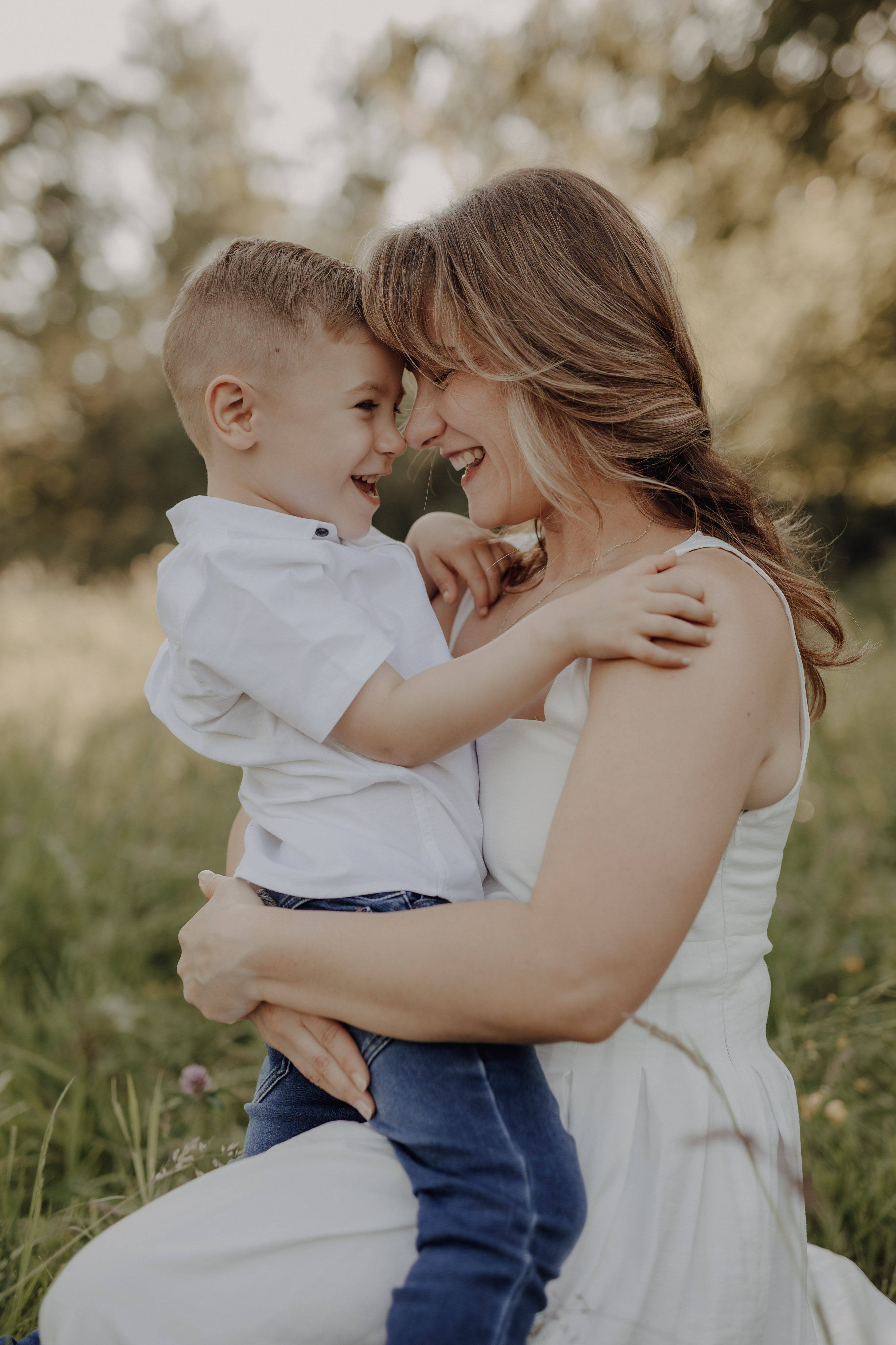 Eine Frau in einem weißen Kleid kniet während eines Familienfotoshootings auf einer Wiese und hält einen kleinen Jungen mit kurzen Haaren, der ein weißes Hemd und Jeans trägt. Sie lächeln, ihre Stirnen berühren sich und fangen einen zarten Moment der Nähe und Freude ein. Im Hintergrund sind Bäume zu sehen.