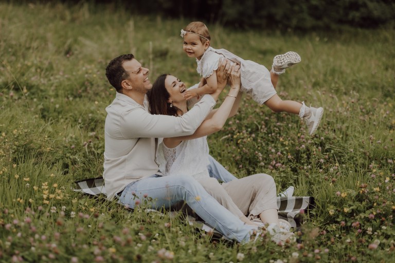 Eine fröhliche Familie genießt ein Picknick auf einer Wiese. Ein Mann, der auf einer Decke sitzt, hebt ein lächelndes kleines Mädchen in die Luft, während die Frau neben ihm das Kind liebevoll stützt. Alle drei, in leichter Freizeitkleidung gekleidet, machen perfekte Familienfotos inmitten von Grün und Wildblumen.