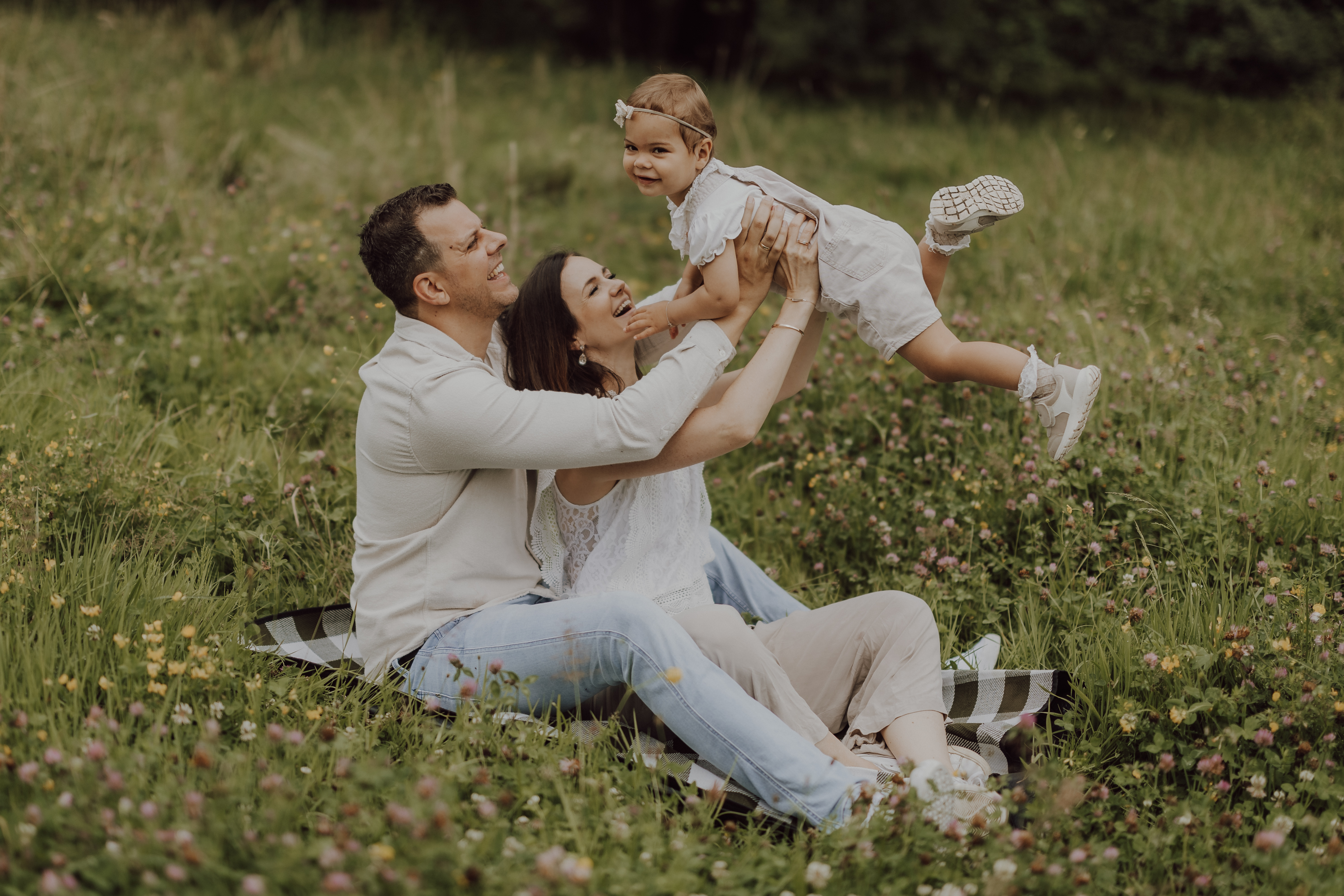 Eine fröhliche Familie genießt ein Picknick auf einer Wiese. Ein Mann, der auf einer Decke sitzt, hebt ein lächelndes kleines Mädchen in die Luft, während die Frau neben ihm das Kind liebevoll stützt. Alle drei, in leichter Freizeitkleidung gekleidet, machen perfekte Familienfotos inmitten von Grün und Wildblumen.