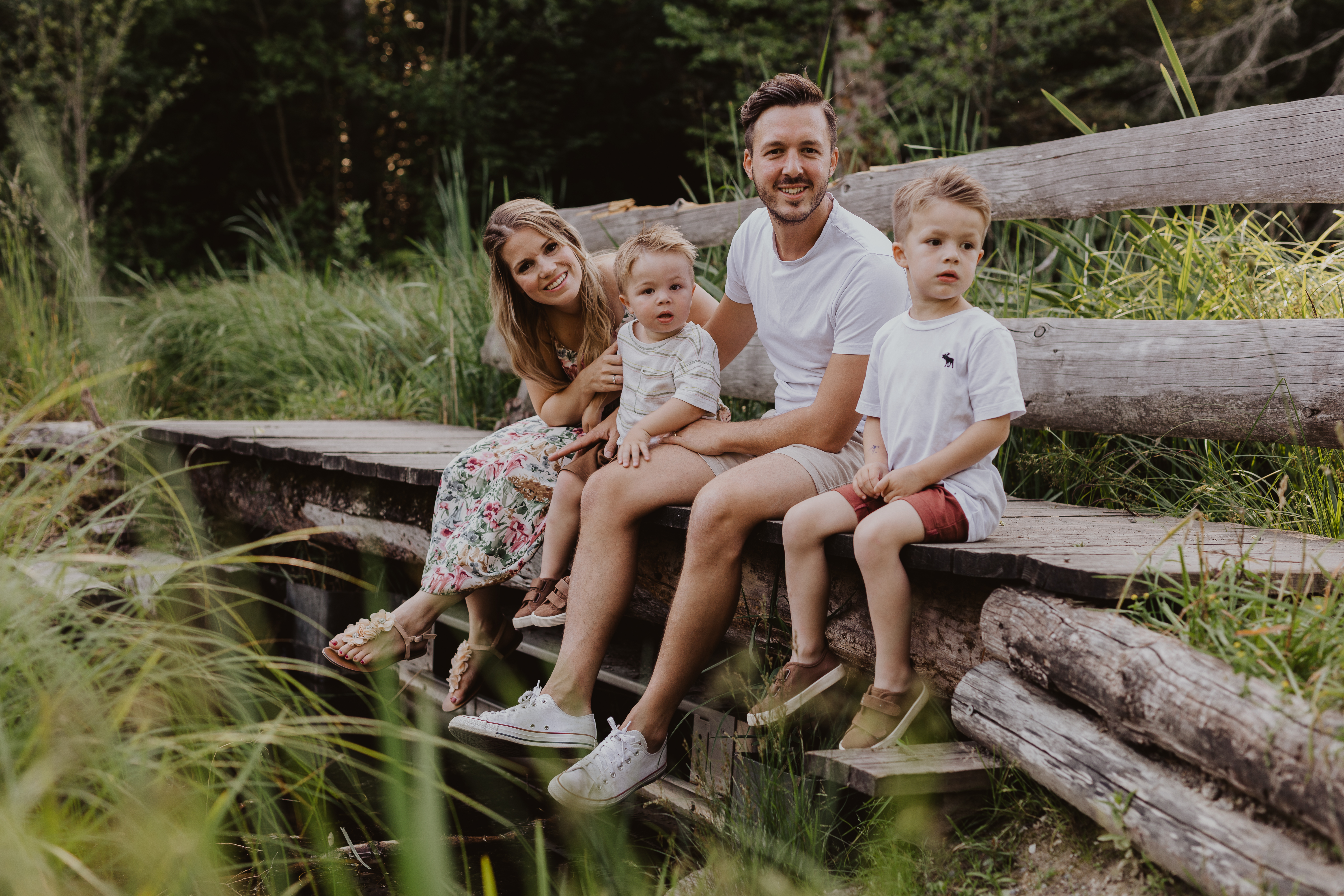 Eine vierköpfige Familie sitzt auf einer kleinen Brücke mitten in der Natur. Sie schauen in die Kamera und lachen fröhlich während des Familienshootings.