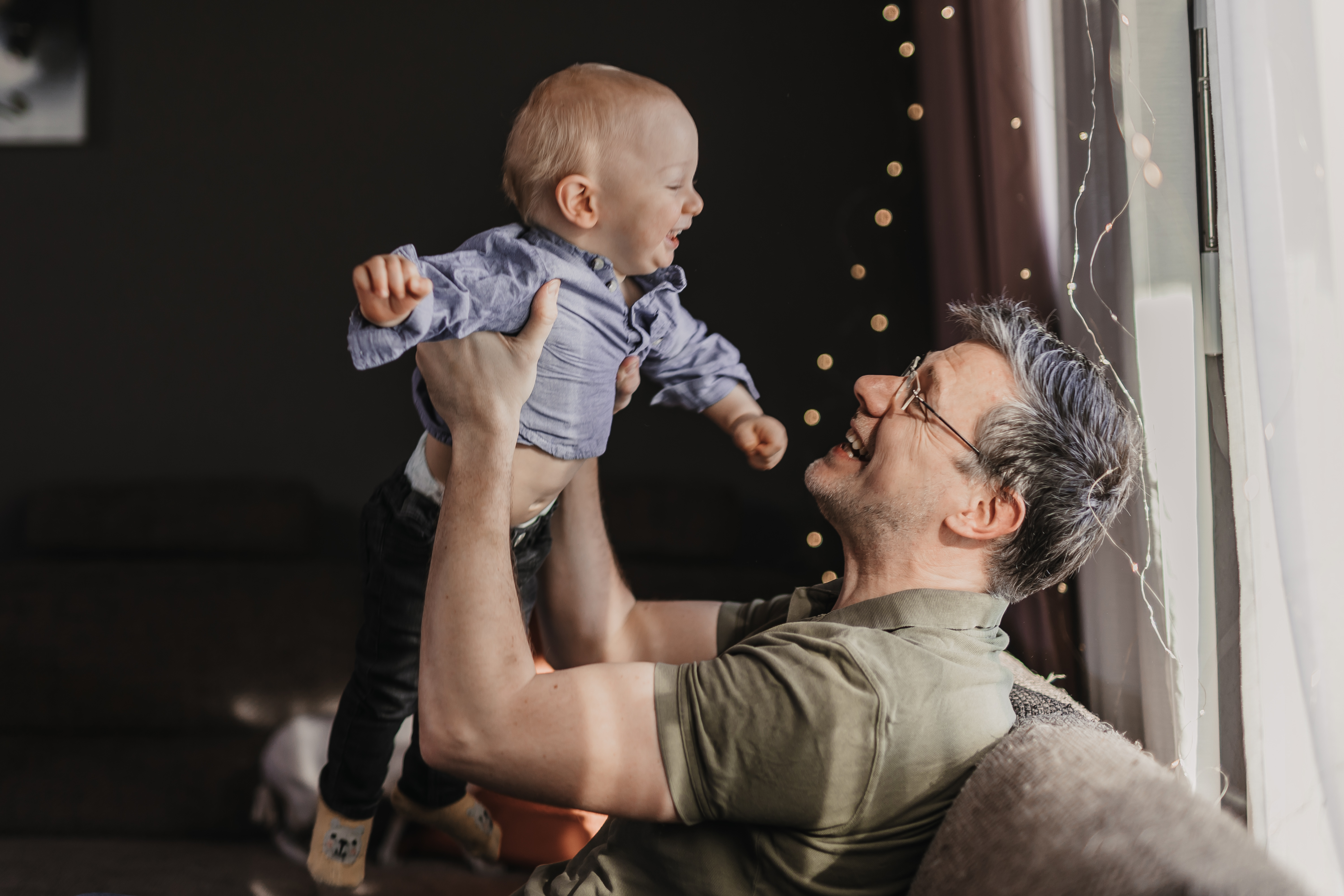 Ein Mann mit Brille und grünem Hemd sitzt lächelnd auf einem Sofa und hebt ein lachendes Baby in blauem Hemd und dunkler Hose in die Luft. Sie stehen beide neben einem Fenster, im Hintergrund schmücken Lichterketten die Lichterkette. Die Szene ist warm beleuchtet und schafft eine gemütliche Atmosphäre, die den Fotoshooting-Service von Tasha Ender Photography perfekt in Szene setzt.