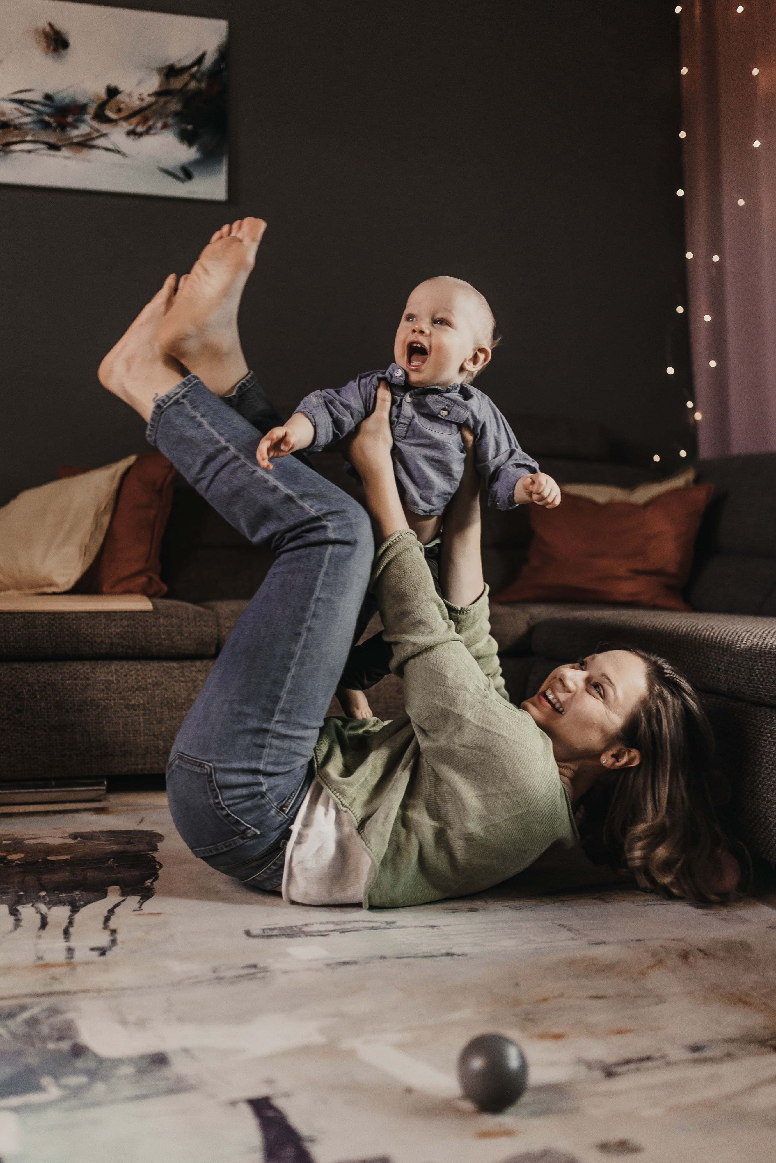 Eine fröhliche Frau spielt mit einem lachenden Baby auf dem Boden eines gemütlichen Wohnzimmers, perfekt für Familienfotos. Die Frau liegt auf dem Rücken und hält das Baby mit den Füßen hoch. Der warm beleuchtete Raum ist mit Lichterketten, einer Couch und einem Gemälde an der Wand dekoriert und bietet somit eine ideale Kulisse für ein Fotoshooting Zuhause.
