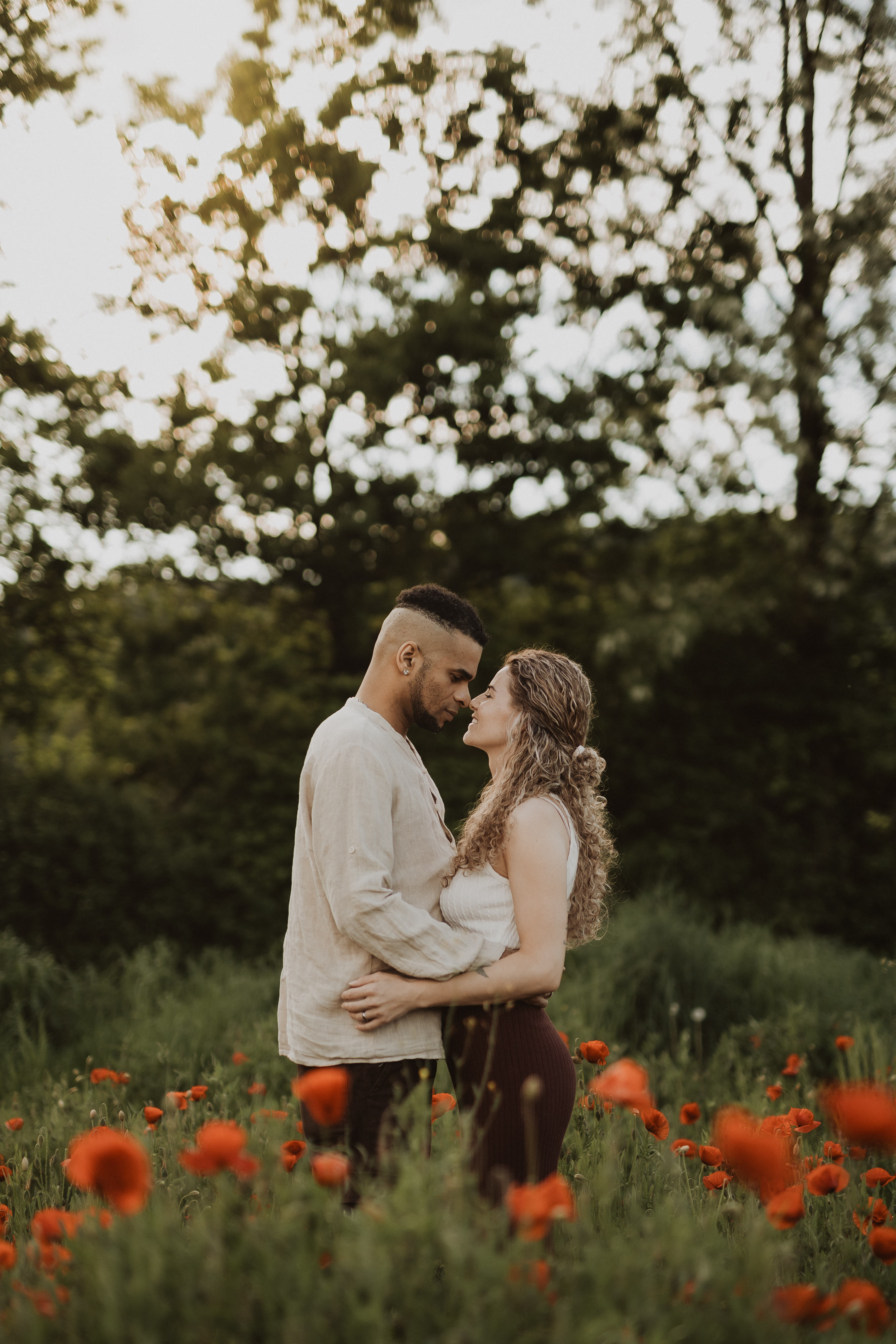 Ein Paar steht in einem Feld aus roten Blumen, einander zugewandt und umarmend, während die Sonne sanft durch die Bäume im Hintergrund scheint. Sie sind von üppigem Grün umgeben, was eine ruhige und intime Atmosphäre schafft, die perfekt für ein Familienshooting zu Hause ist.