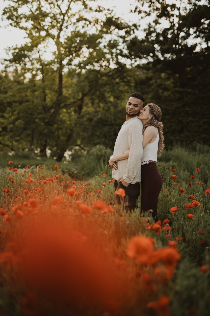 Ein Paar umarmt sich in einem Feld roter Mohnblumen während eines Fotoshootings in der Natur. Der Mann im hellen Hemd steht nach vorne gerichtet, während die Frau in weißem Oberteil und dunkler Hose hinter ihm steht und ihre Arme um seine Brust gelegt hat. Hohe, üppige Bäume bilden den Hintergrund.