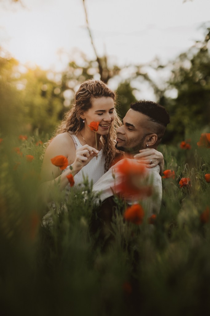 Ein Paar sitzt eng beieinander in einem Feld aus roten Blumen und teilt einen zärtlichen Moment. Die Frau hält eine Blume nah an das Gesicht des Mannes, beide lächeln und blicken sich liebevoll an. Um sie herum sind üppige grüne Pflanzen und ein sanft beleuchteter, ruhiger Hintergrund – ein wahrer Ausdruck der Essenz eines romantischen Fotoshootings in der Natur.