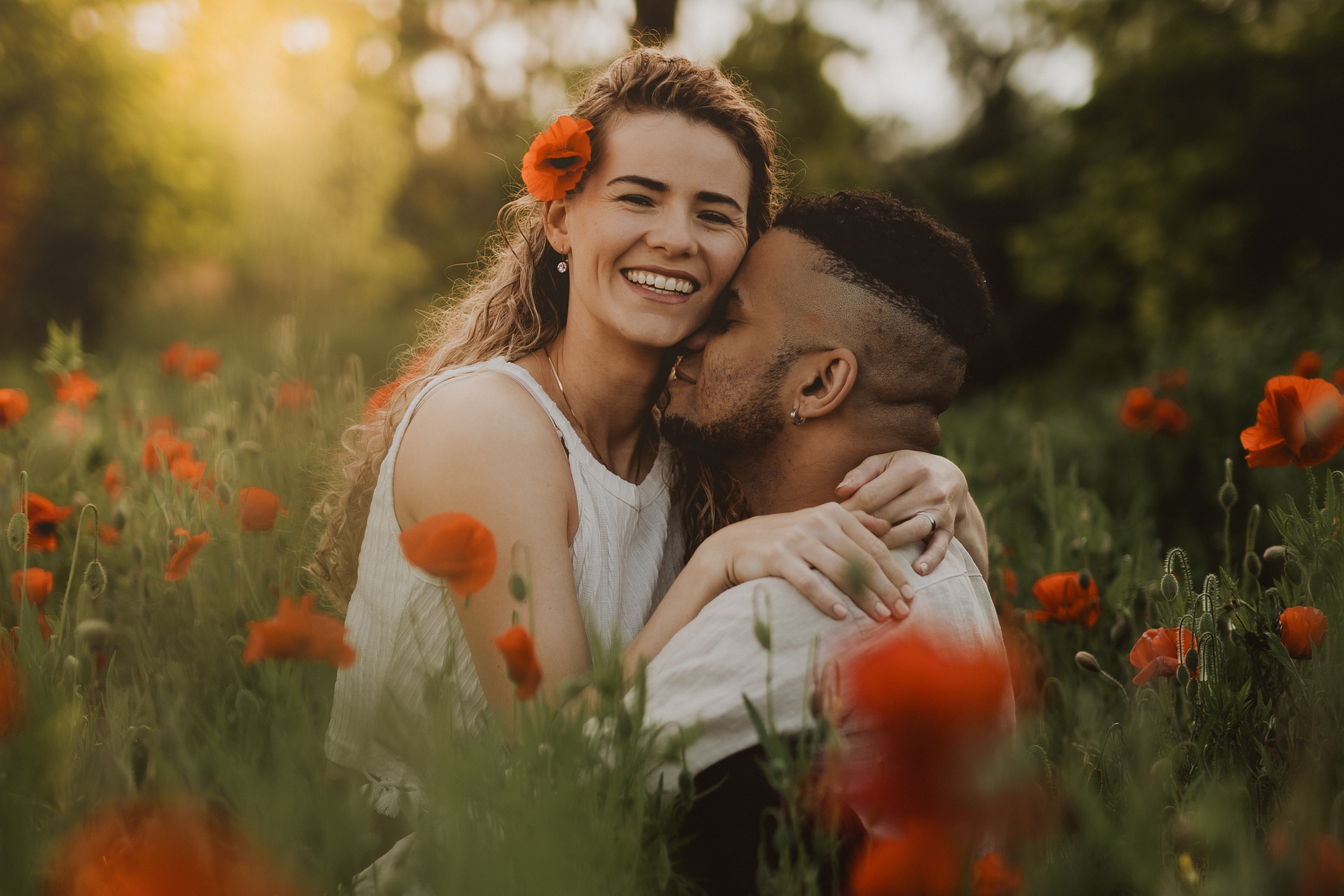 Ein Paar umarmt sich während eines Fotoshootings in der Natur in einem Feld roter Mohnblumen. Die Frau mit lockigem Haar und einer roten Blume hinter dem Ohr lächelt strahlend, während der Mann mit kurzen Haaren sie auf die Wange küsst. Die Sonne wirft einen warmen Schein auf die Szene und unterstreicht ihre glücklichen Ausdrücke.