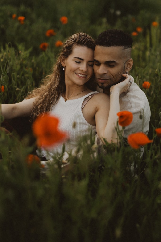 Ein Paar sitzt eng beieinander in einem Feld mit hohem Gras und leuchtend roten Mohnblumen. Die Frau mit den lockigen Haaren lehnt sich liebevoll an den Mann, der kurze Haare und einen gestutzten Bart hat. Sie wirken entspannt und glücklich während ihres Fotoshootings, umgeben von der natürlichen Schönheit der Blumen.