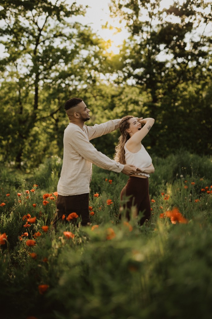 Ein Mann und eine Frau stehen in einem Feld mit hohem Gras und roten Blumen. Der Mann, der ein beiges Langarmhemd trägt, streckt seinen Arm in Richtung der Frau aus. Die Frau, die ein weißes ärmelloses Oberteil trägt, berührt mit der linken Hand ihren Kopf und schaut weg. Bäume und Sonnenlicht bilden die perfekte Kulisse für natürliche Familienfotos.