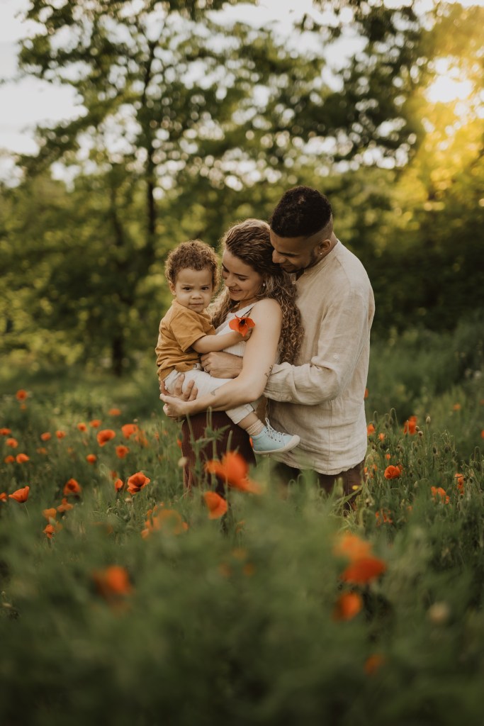 Eine dreiköpfige Familie steht in einem sonnenbeschienenen Feld aus orangefarbenen Blumen. Die Mutter hält ihr kleines Kind, das die Blumen anlächelt, während der Vater hinter ihnen steht und sanft seine Hände auf die Schultern seiner Partnerin legt. Sie alle teilen einen zärtlichen Moment inmitten des üppigen Grüns während eines natürlichen Familienshootings.