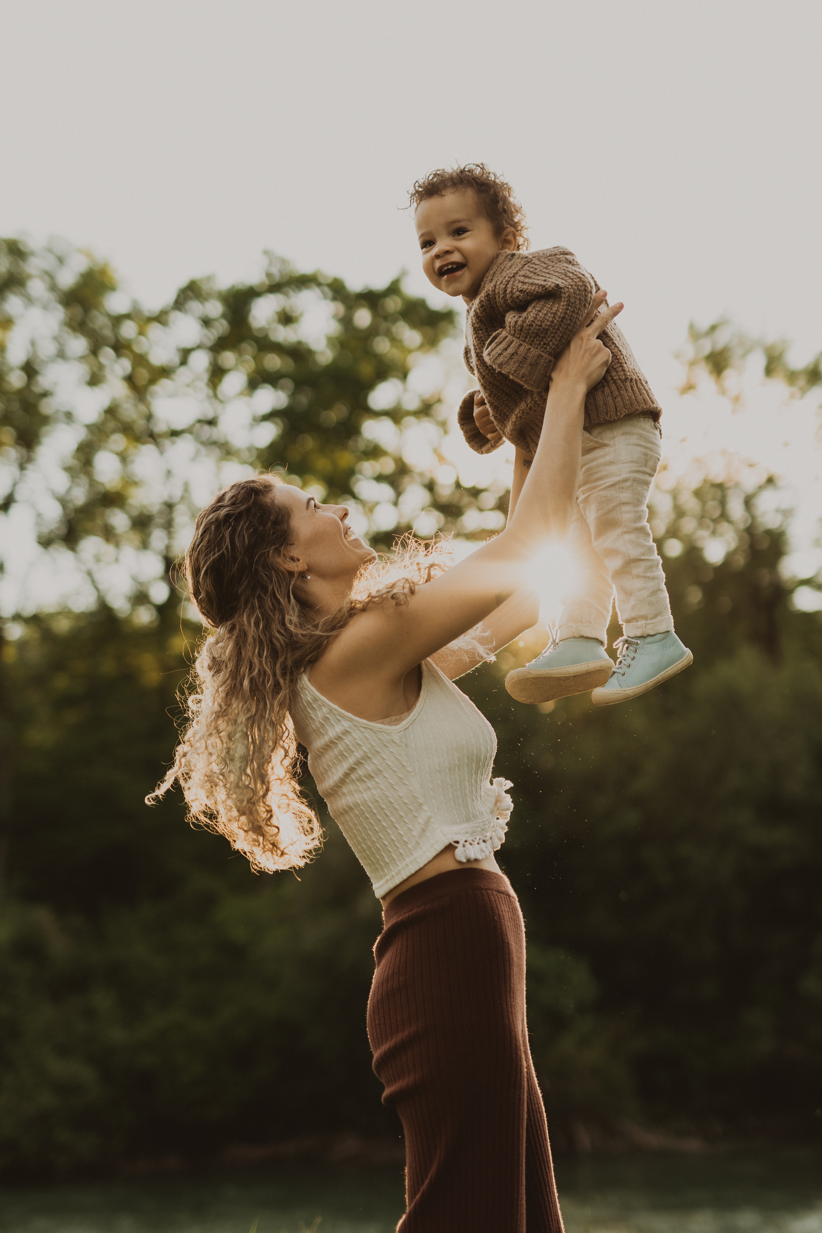 Eine Frau mit lockigem Haar und einem ärmellosen weißen Oberteil hebt während eines Fotoshootings in der Natur ein lächelndes Kleinkind in die Luft. Die Sonne beleuchtet sie von hinten und grünes Laub trägt zu dem warmen und freudigen Moment bei. Das Kleinkind, bekleidet mit einem braunen Pullover und blauen Schuhen, vervollständigt diese Szene natürlicher Familienfotos.