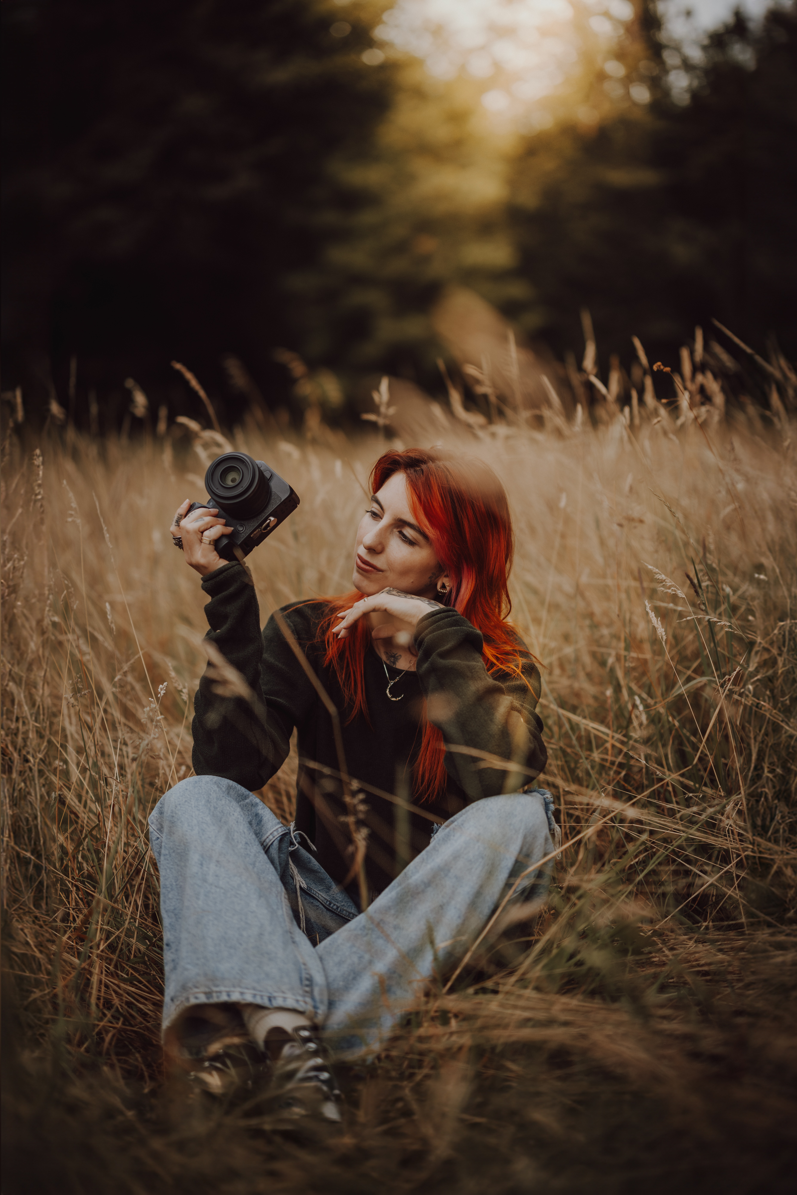 Eine Person mit langen roten Haaren sitzt in einem Feld mit hohem Gras und hält eine Kamera in der Hand. Lässig gekleidet in einem dunklen Pullover und hellblauen Jeans blickt sie mit nachdenklichem Gesichtsausdruck in die Kamera. Der Hintergrund ist verschwommen, Sonnenlicht scheint durch die Bäume und fängt die Essenz eines Aargauer Fotoshootings in der Nähe von Baden ein.