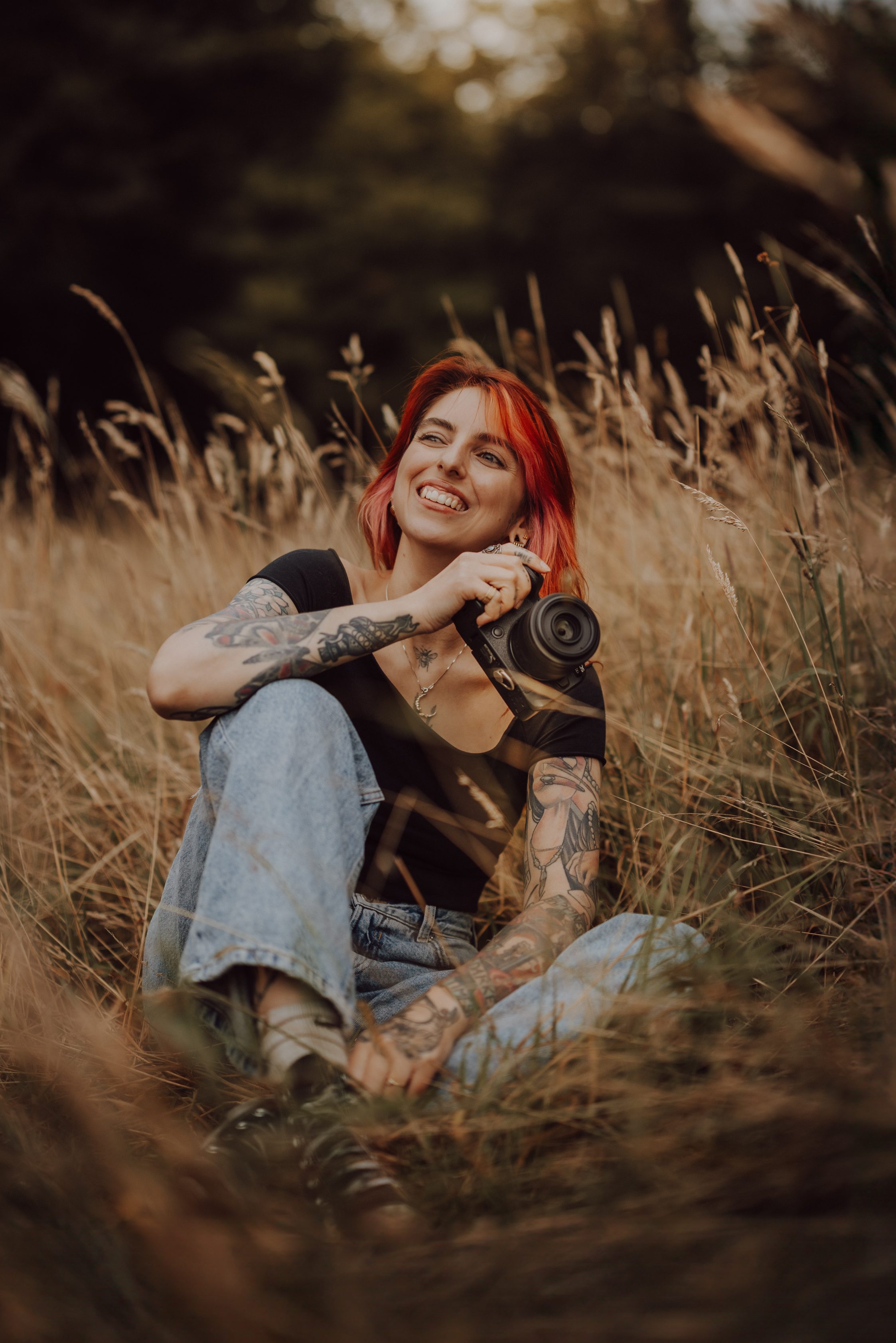 Eine Person mit roten Haaren, Tattoos und einem schwarzen Hemd sitzt lächelnd auf einer Wiese im Aargau und hält eine Kamera in der Hand. Sie hat ein Knie hochgezogen und ist von hohem Gras umgeben, was darauf hindeutet, dass sie ein Fotoshooting in dieser natürlichen Umgebung im Freien genießt.